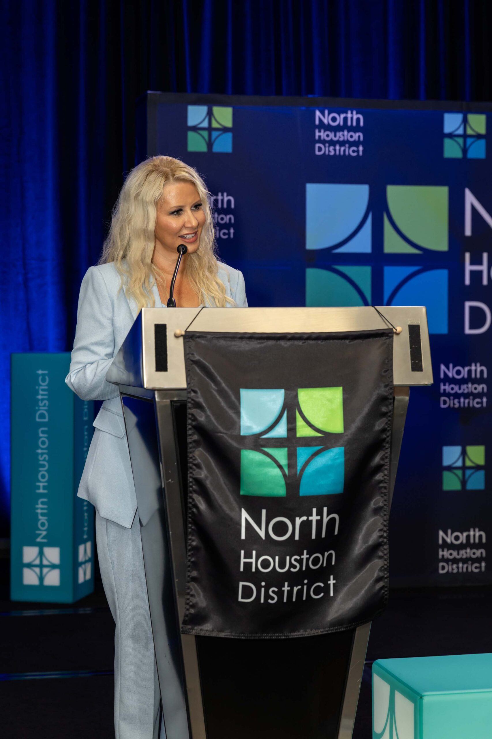 A woman in a light blue suit speaks at a podium with a North Houston District banner.