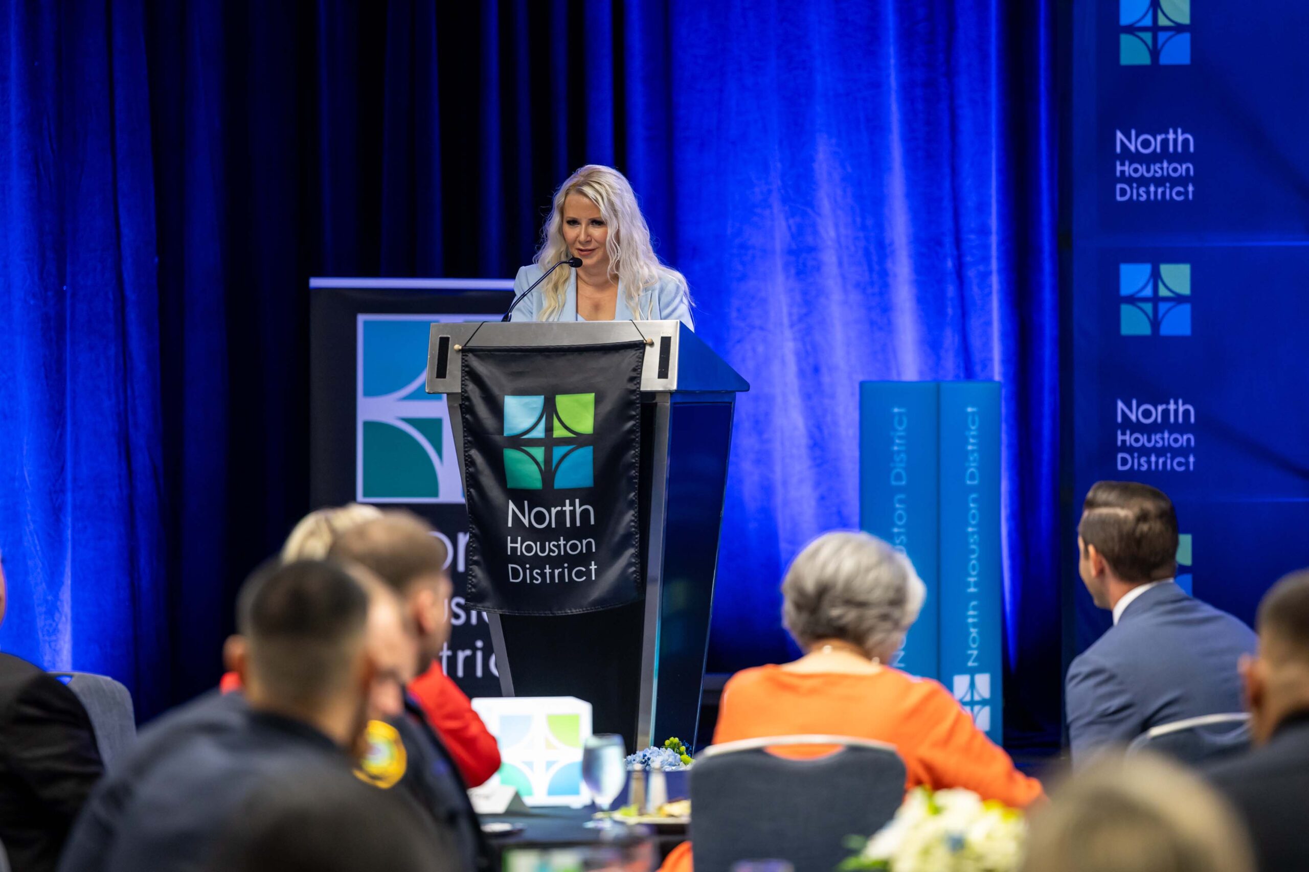 A woman in a light blue suit stands behind a podium addressing the crowd at the awards luncheon.