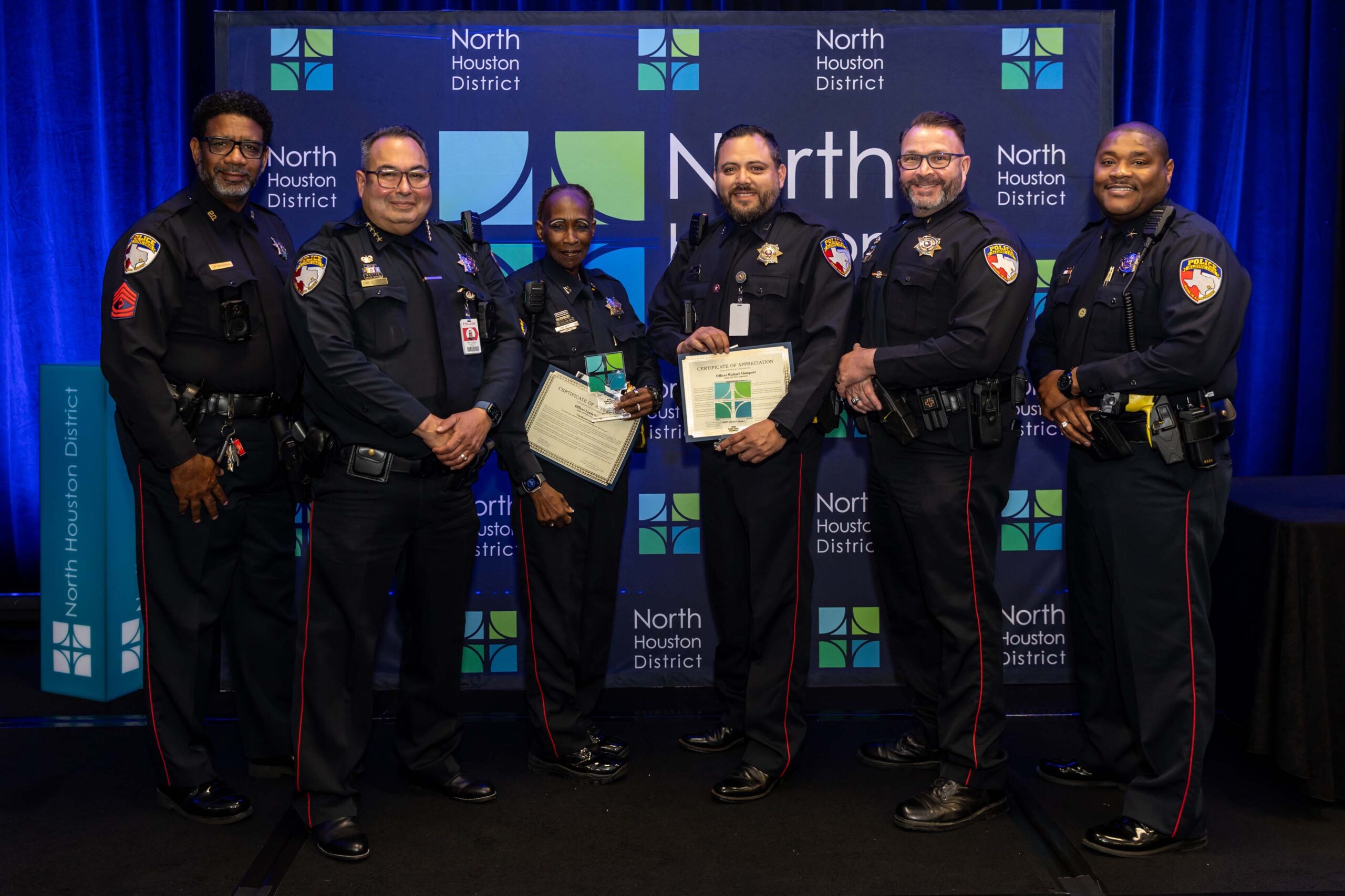 Six officers from a Aldine ISD Police Department stand in a row, with two officers holding certificates in front of a North Houston District banner.