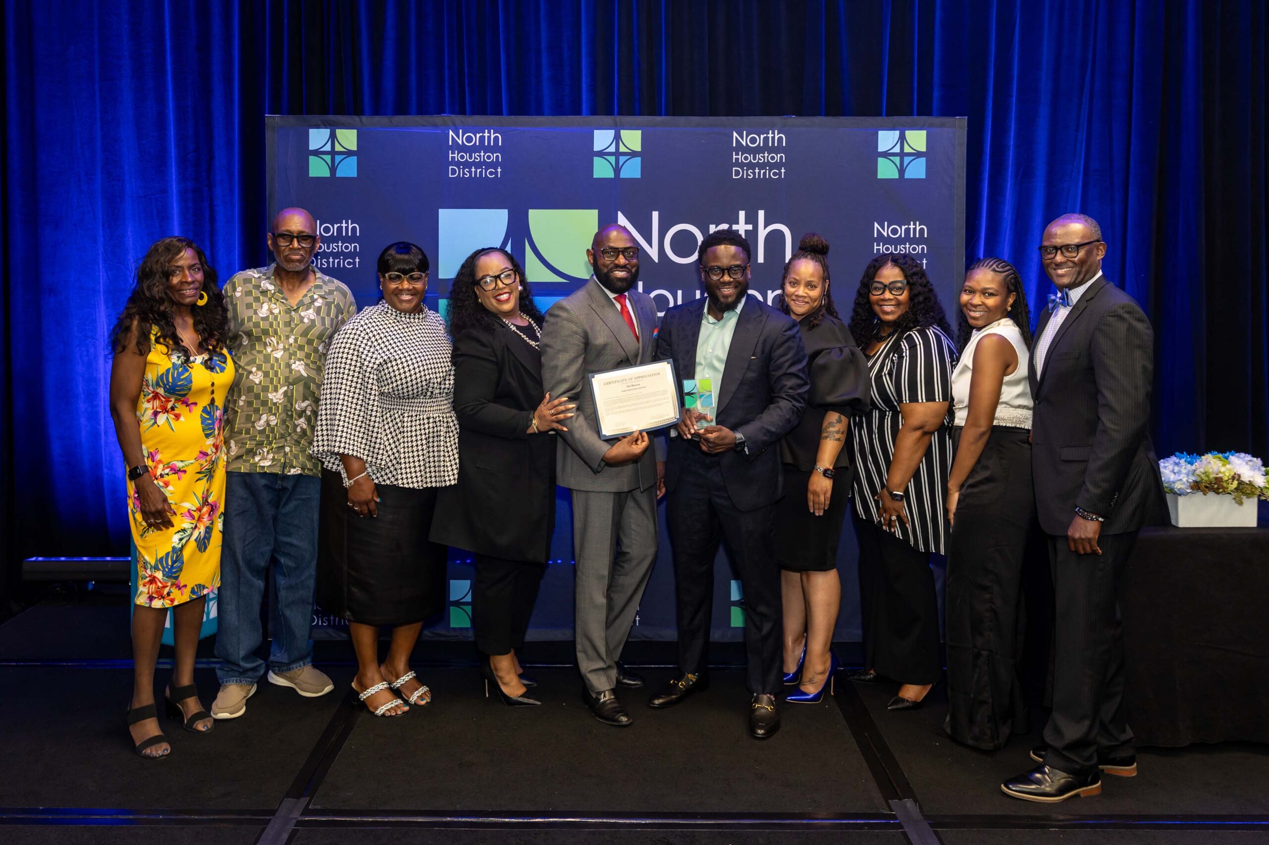 A group of 11 people pose on stage with certificates and awards at the Public Safety Awards event, smiling in front of the North Houston District backdrop.