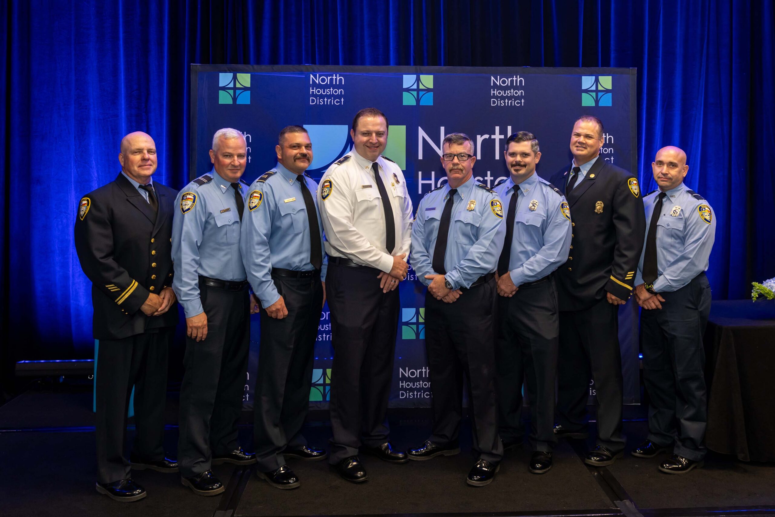 Eight uniformed officers from a fire department pose for a group photo in front of a North Houston District backdrop.