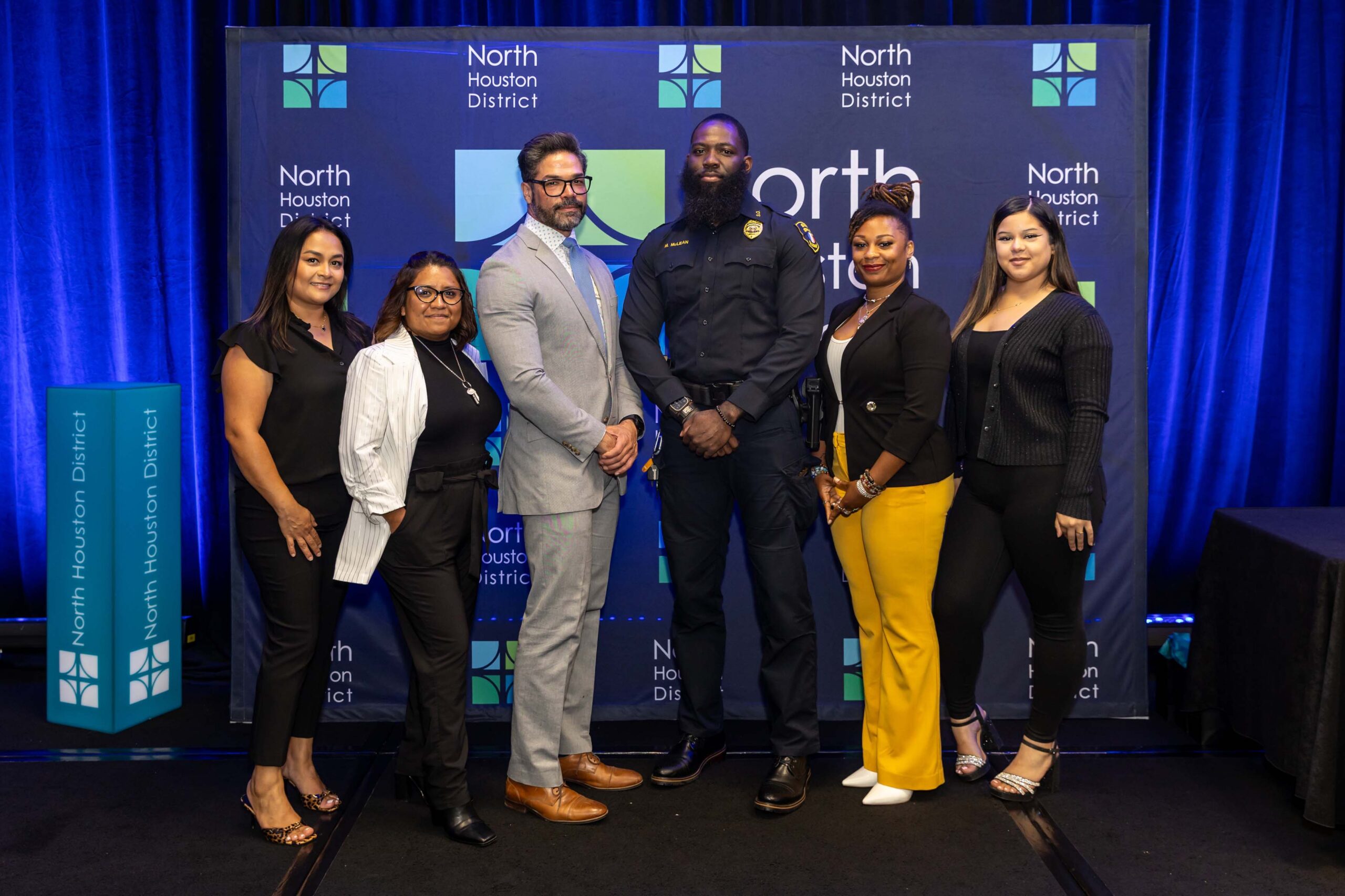 Six individuals pose together in front of a North Houston District backdrop. One is in a dark police uniform, while the others wear professional or business casual attire.