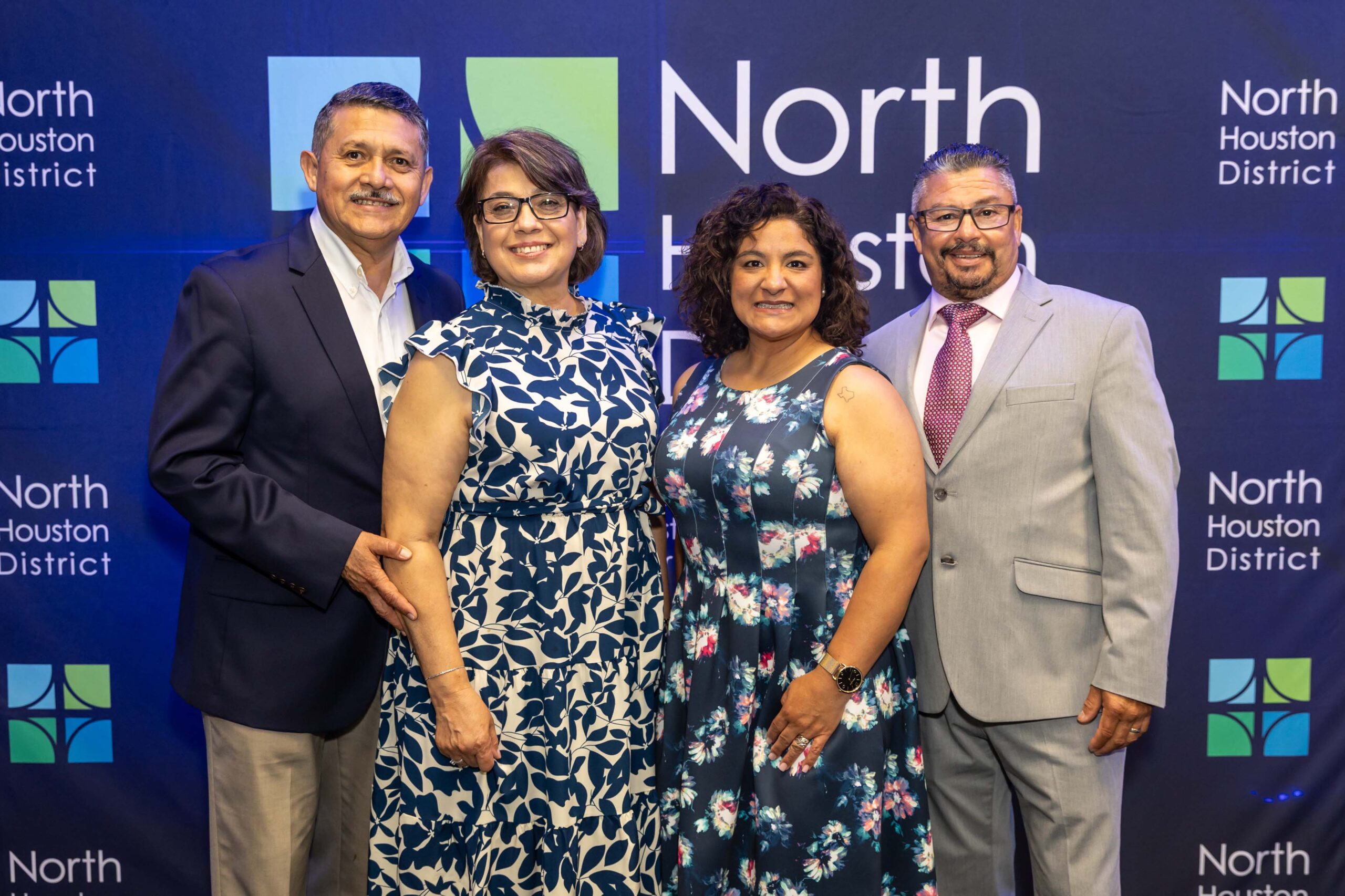 Two men and two women in floral and professional outfits smile together in front of the North Houston District step-and-repeat.