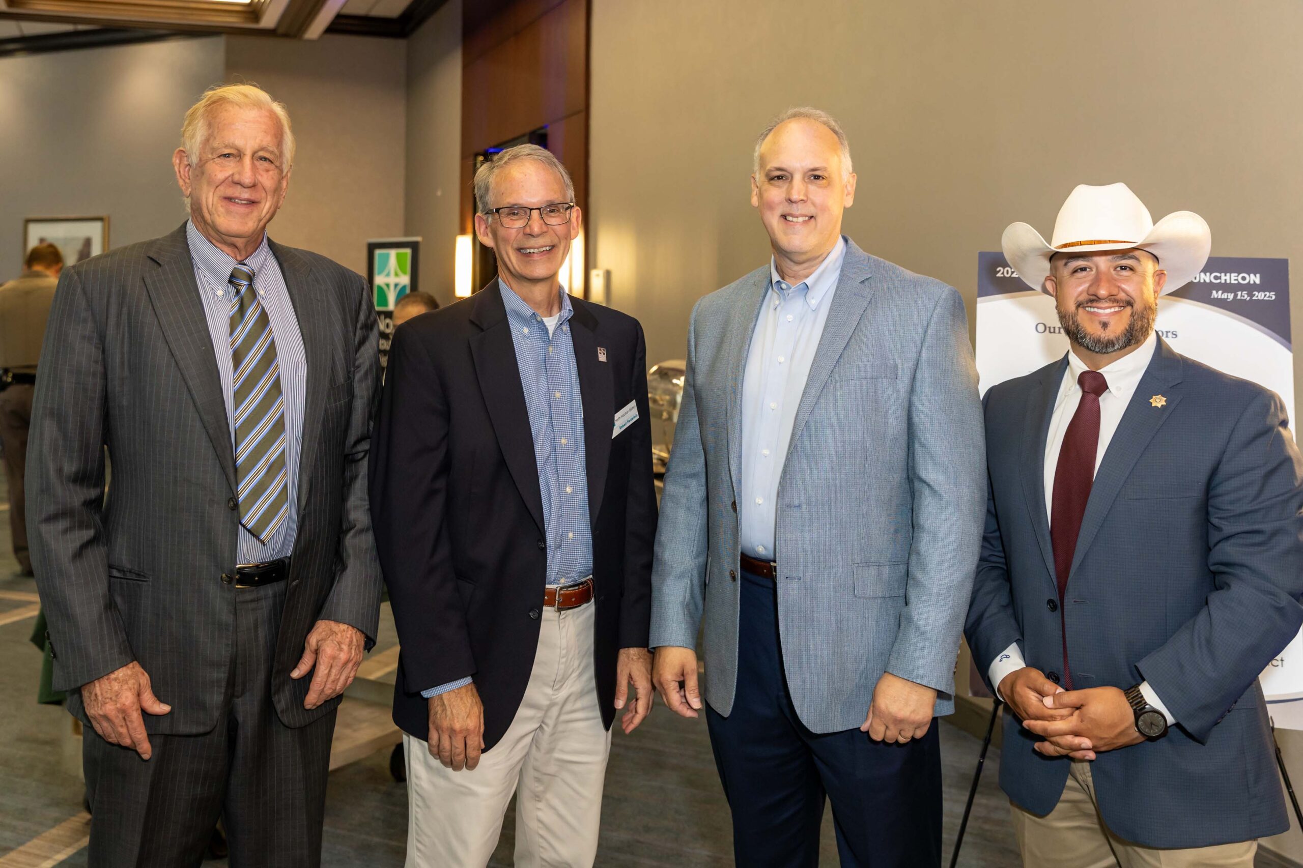 Four men stand in the lobby, smiling at the camera. One wears a white cowboy hat; others wear suits or blazers in shades of gray and blue.