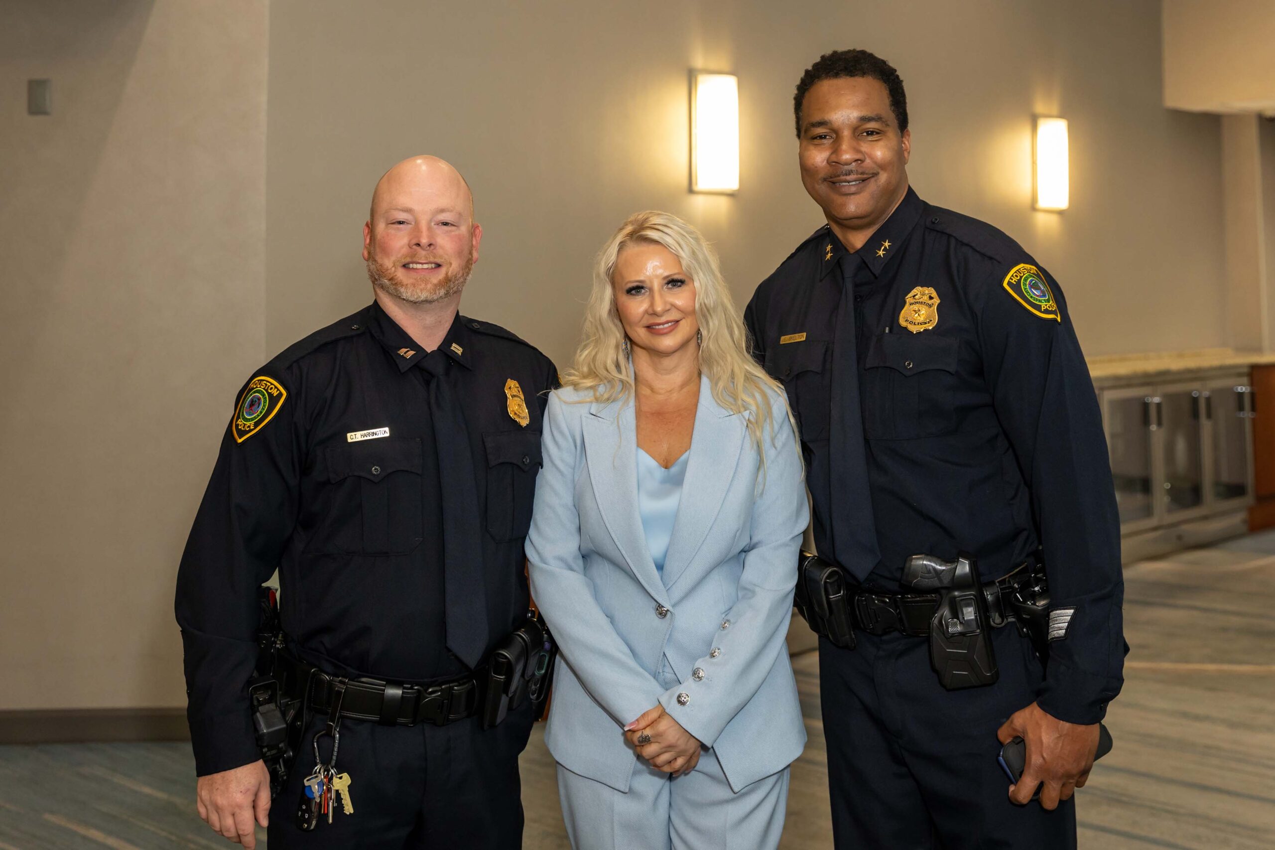 Two uniformed Houston Police Department officers stand on either side of a woman in a light blue suit with long blond hair.