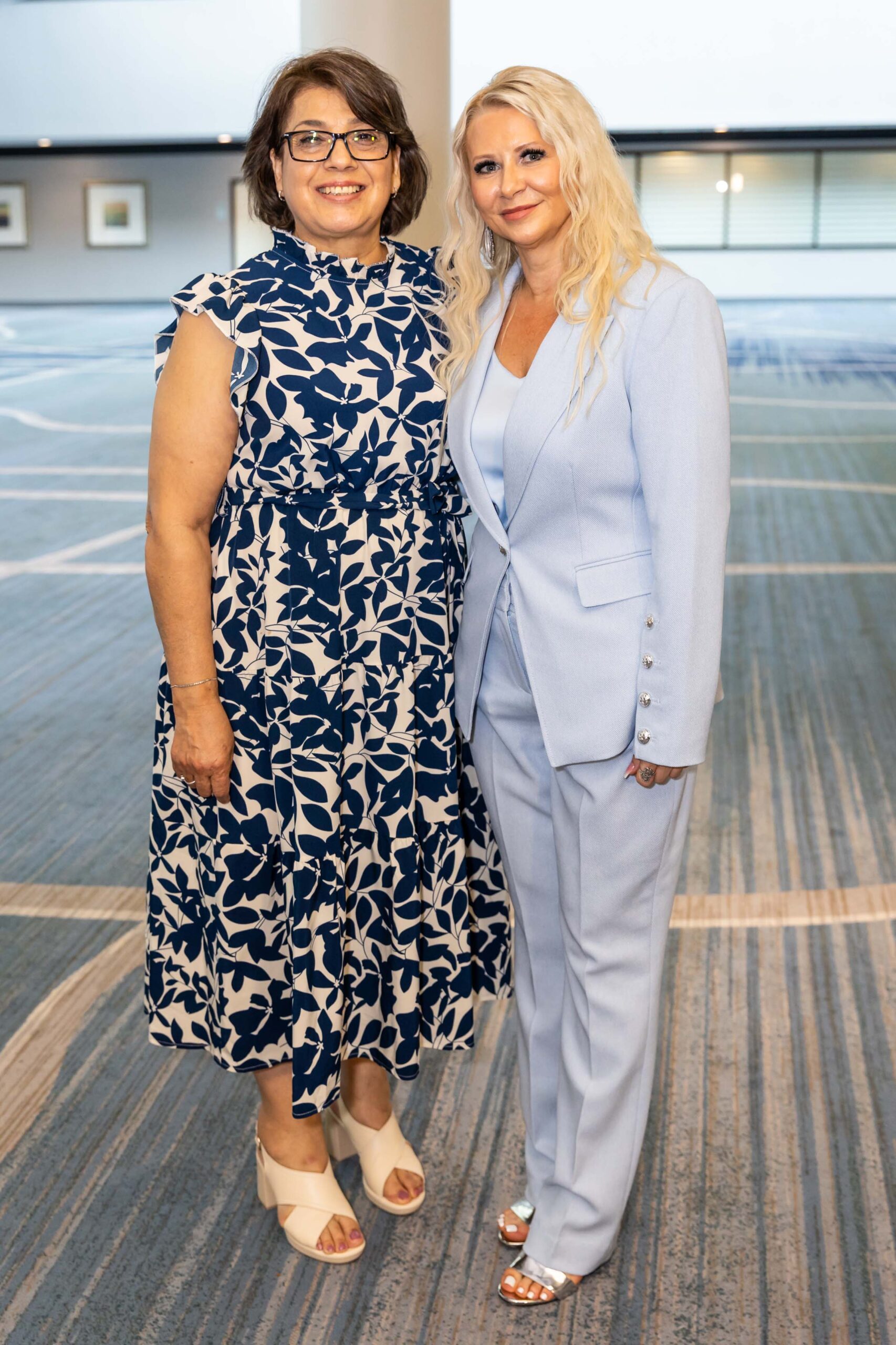 Two women stand side by side smiling; one wears a sleeveless navy floral dress, the other a powder blue pantsuit.