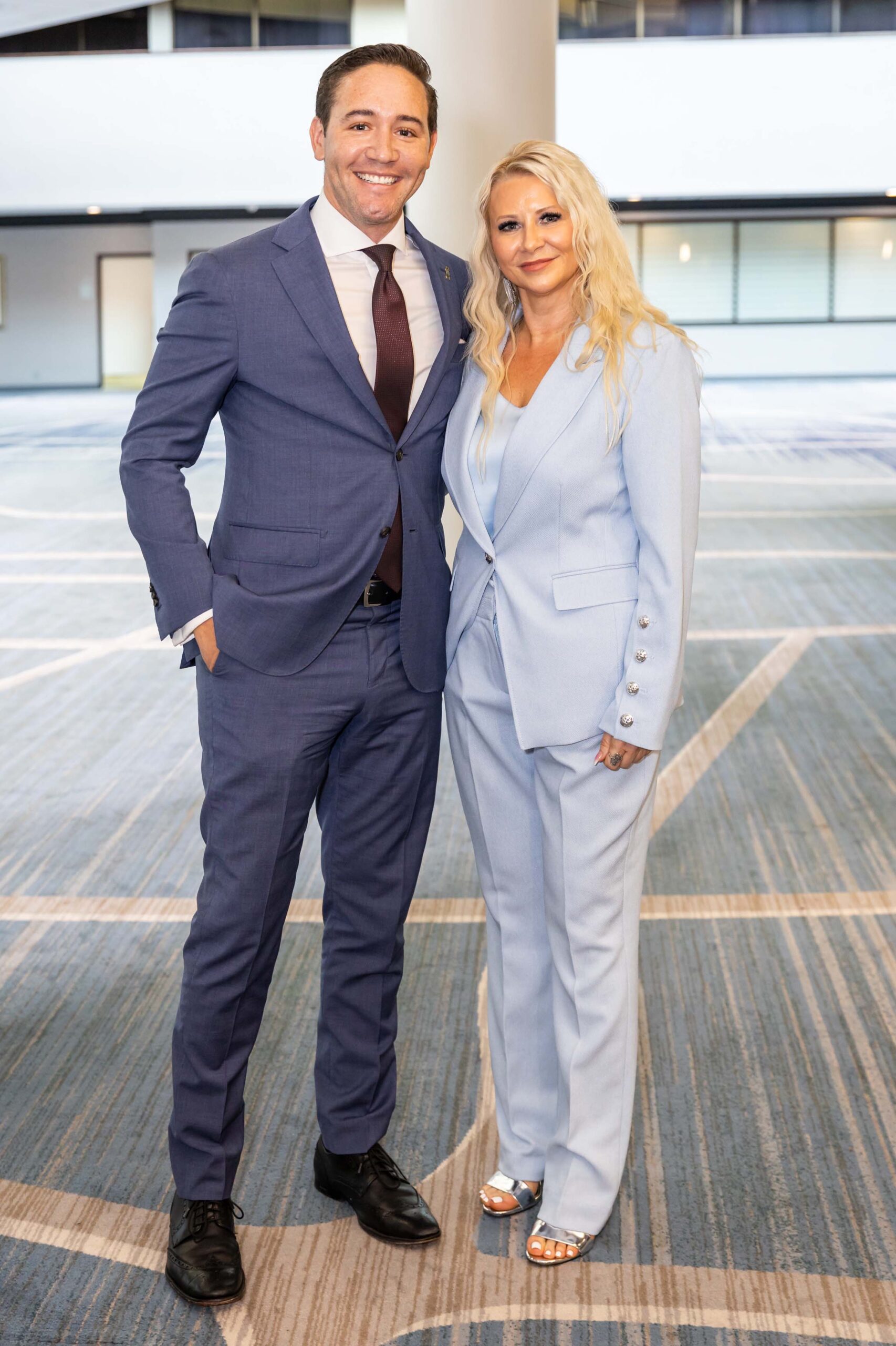 A man in a blue suit stands next to a woman in a light blue pantsuit, both smiling in a well-lit indoor space.