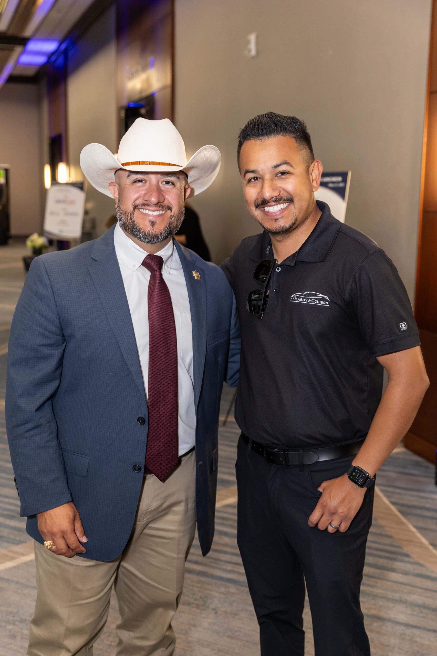 Two smiling men pose together; one wears a white cowboy hat and blue blazer.