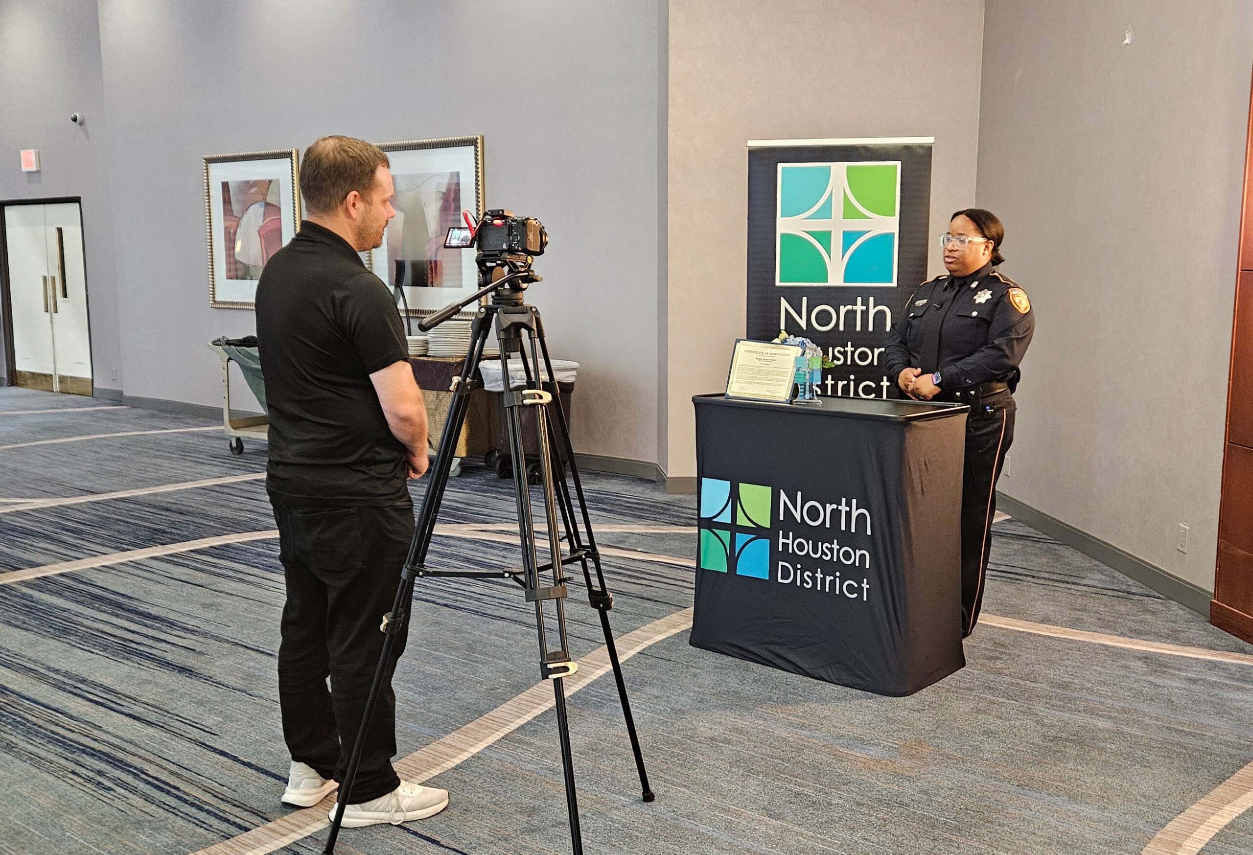A uniformed officer is interviewed on camera beside a District backdrop and award table.