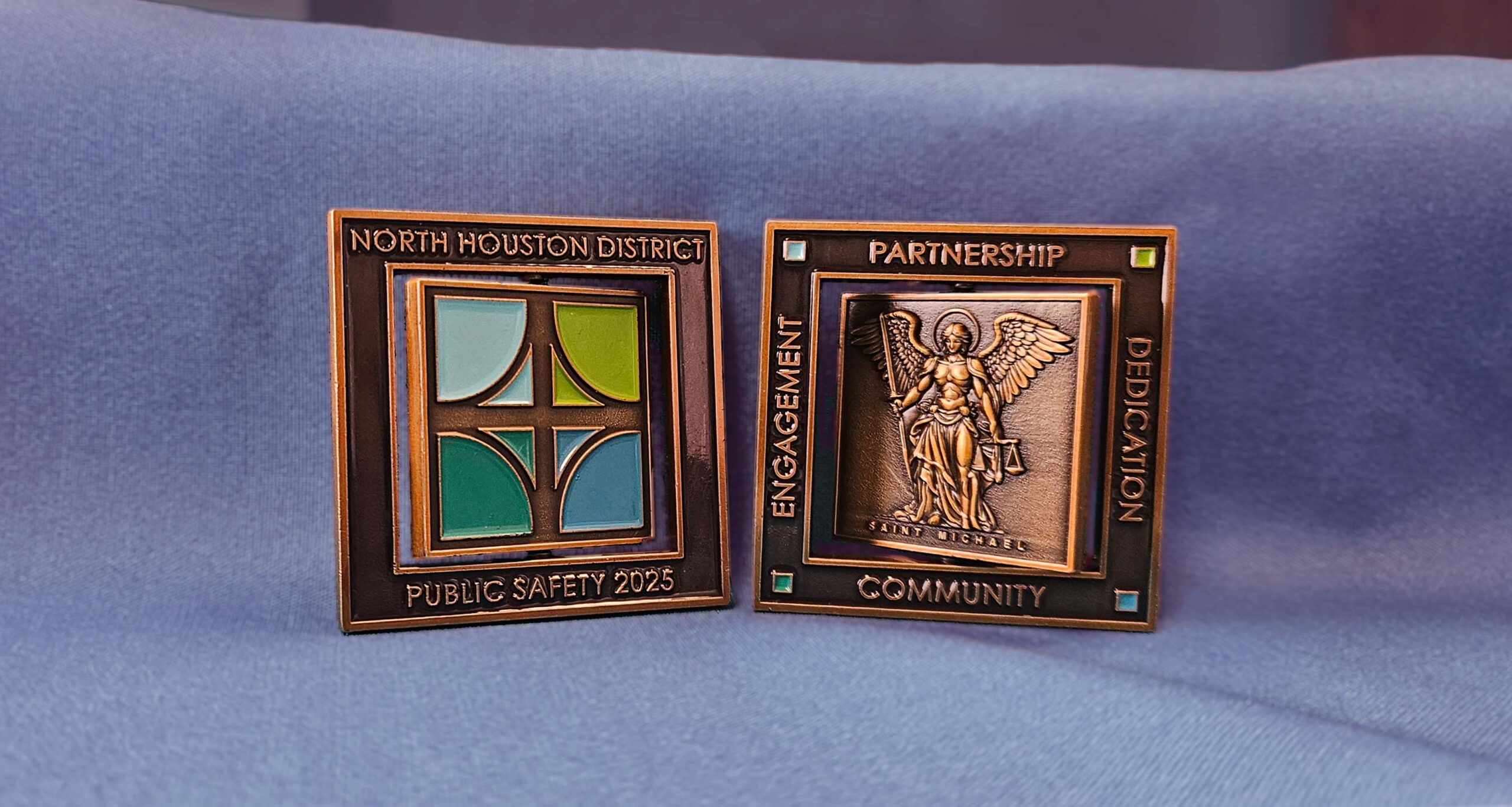 The 2025 Commemorative Challenge Coin rest on blue fabric. One side features the North Houston District logo; the other depicts Saint Michael and the words Partnership, Engagement, Dedication, and Community.