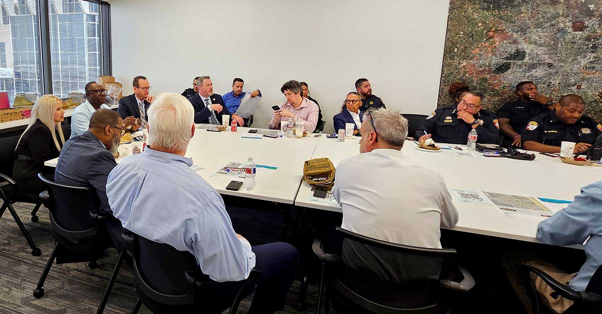 Meeting attendees listen intently during a Public Safety Committee session, with law enforcement and civic leaders seated around a white conference table.