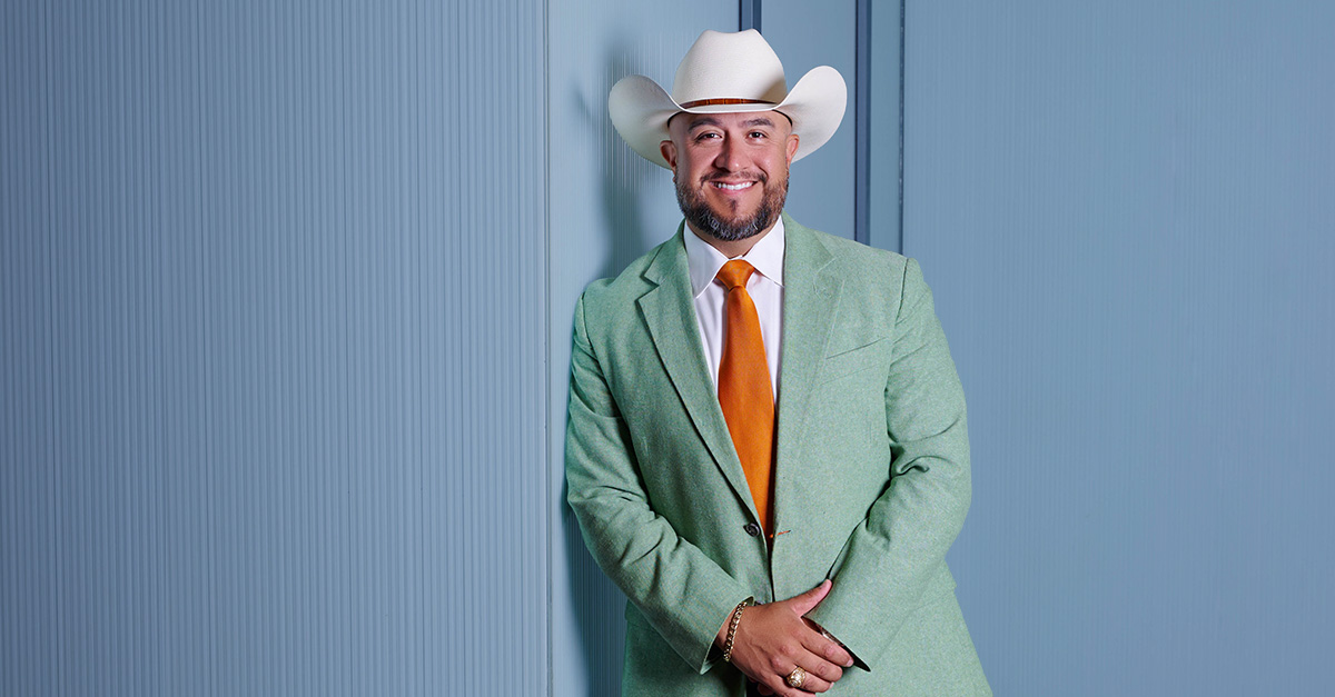 Portrait of a man wearing a light green jacket, orange tie and white cowboy hat, standing against a blue paneled wall.