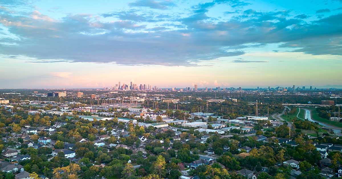 A wide aerial view of Houston showing suburban neighborhoods in the foreground, with the downtown skyline on the horizon under a partly cloudy sky.