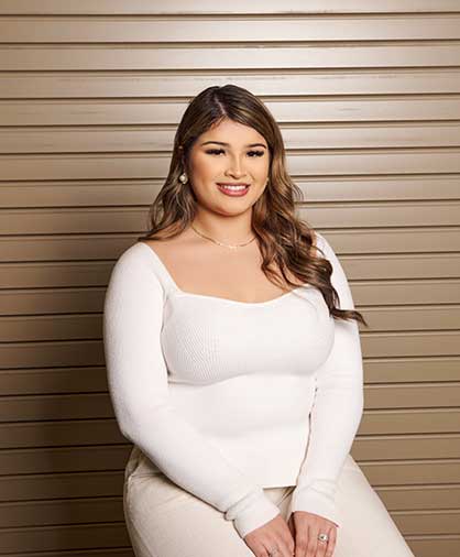 A young woman in a white blouse and light pants sits smiling in front of a tan wood-paneled wall.