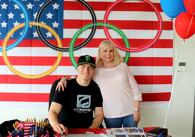 Sally and Connor Fields Two people pose in front of an American flag and Olympic rings display at an event table decorated with patriotic items and balloons.