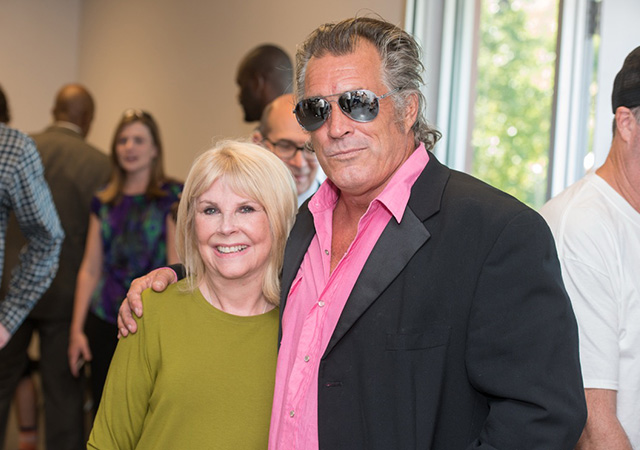 Sally and Steve Olson Two attendees pose together at an indoor event, smiling for the camera while other guests gather in the background.