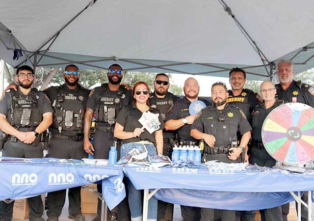 A group of law enforcement officers and community members standing under a tent at National Night Out 2024. The table in front displays items, including bottled water and a spinning prize wheel.