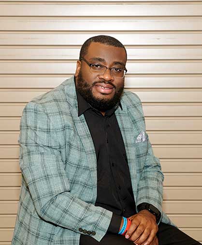 Shawn Bingham Headshot 2 A man in glasses and a green plaid jacket sits against a tan background, leaning slightly forward and smiling, wearing bracelets and a watch.