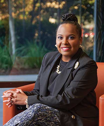 A professional headshot of Sorita Alexander sitting in an orange chair, wearing a black blazer with pearl accessories. She smiles warmly with greenery visible through a window behind her.