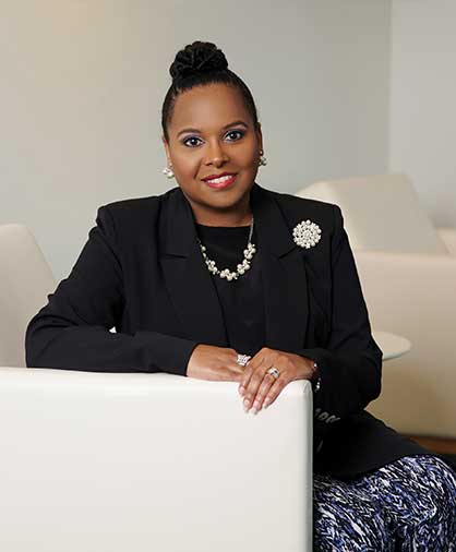 Sorita Alexander poses for a professional headshot, leaning on a white couch. She is dressed in a black blazer, pearl accessories, and a patterned skirt, smiling confidently in a modern office setting.