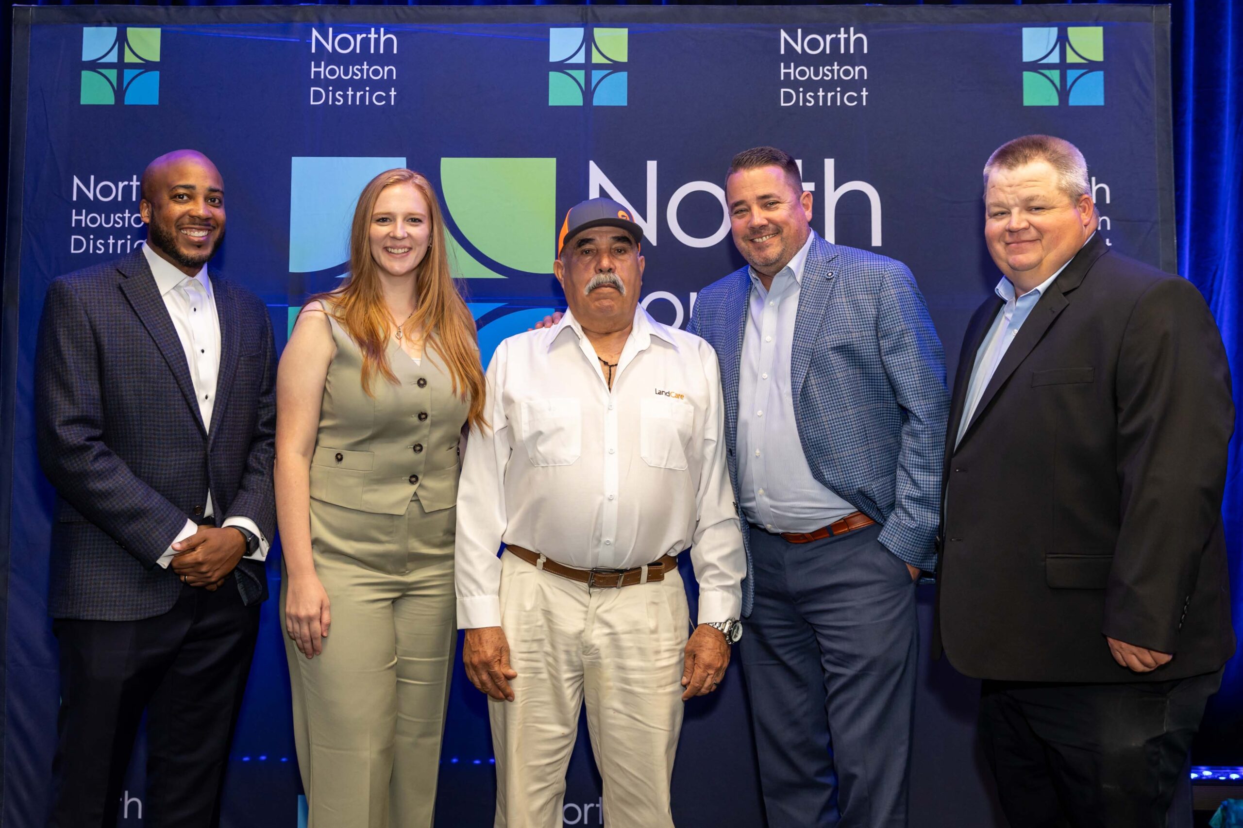 Five people from LandCare, a gold sponsor of the event, pose together in front of the North Houston District banner. One wears a white uniform shirt with the company logo.