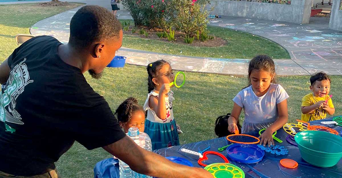 Summer Events Newsroom Graphic A group of young children and an adult engage in a bubble-blowing activity at a park. Colorful bubble wands and bowls cover the table, and chalk drawings are visible on the sidewalk in the background.