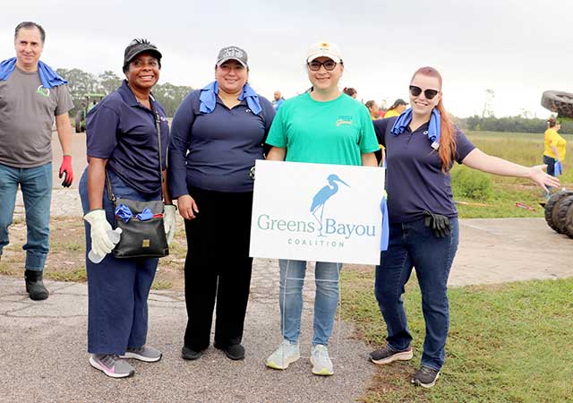 Tina Poindexter stands with four other volunteers outdoors holding a Greens Bayou Coalition sign at a community event, all dressed casually and smiling for the photo.