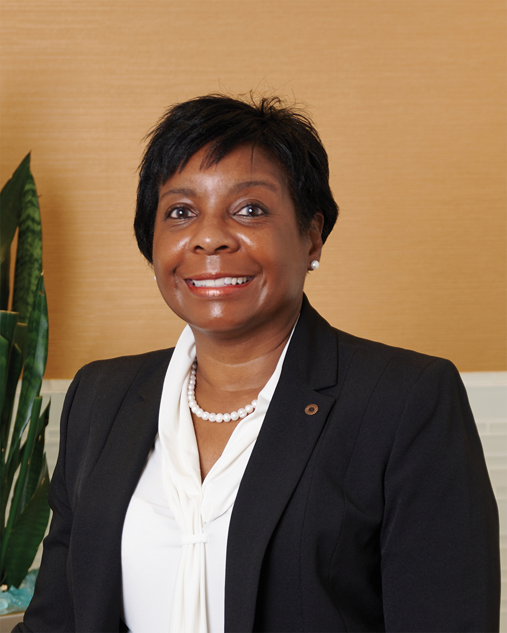 Headshot of Tina Poindexter smiling, wearing a dark blazer and white blouse with a pearl necklace, posed against a tan background.