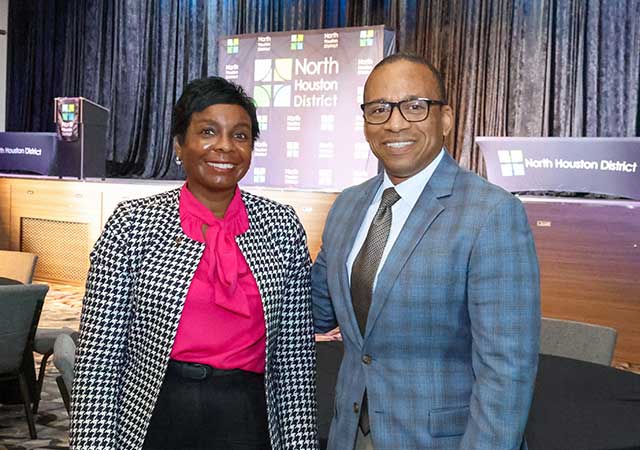 Tina Poindexter and a man in a blue plaid suit stand smiling in front of a stage with North Houston District signage and podiums in the background.