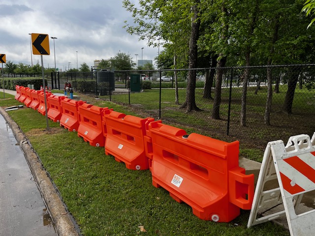 Orange temporary barricades stretch along a grassy area near a curved road. Speed caution signs are visible in the background, and a fence separates the road from Rockstar Energy Bike Park. Trees and overcast skies frame the scene.