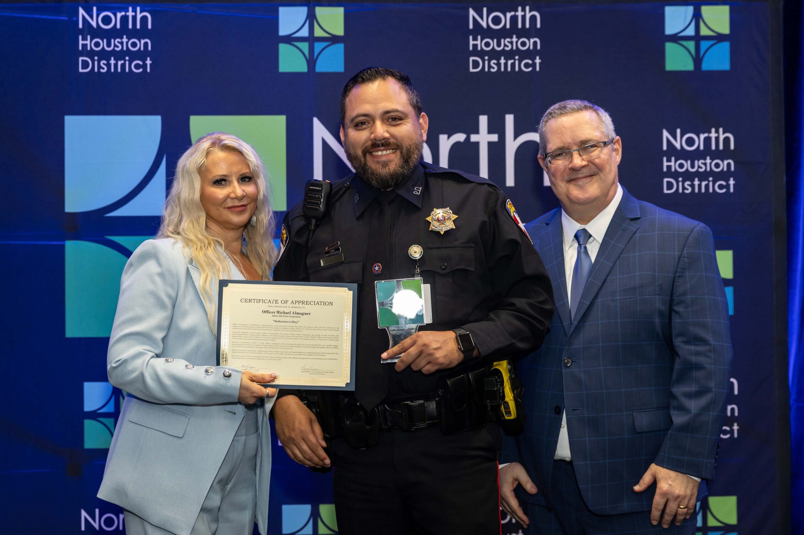 Three people pose together on stage at the North Houston District Public Safety Awards. The woman on the left, wearing a light blue suit, holds a certificate of appreciation. The uniformed officer in the center holds an award. A man in a blue suit stands to the right.