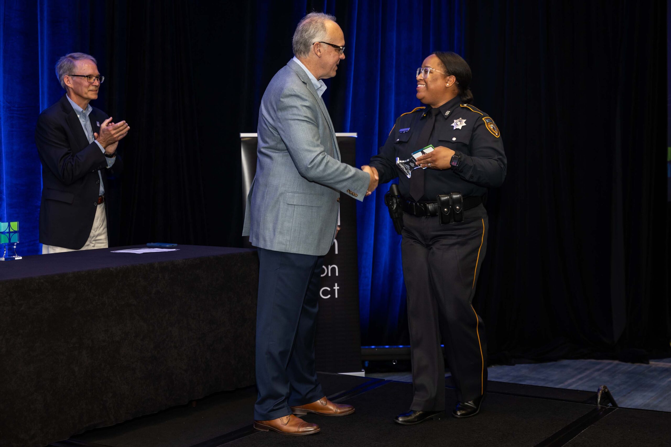 A man in a gray jacket shakes hands with a uniformed female deputy receiving an award on stage. Another man in a navy blazer stands to the left applauding.