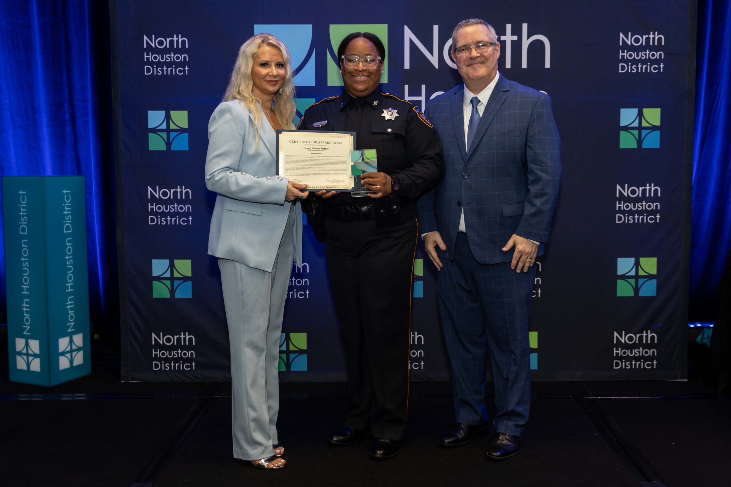 Three people stand on stage smiling. A woman in a light blue suit holds a certificate while a female officer in uniform holds an award. A man in a blue suit stands beside them.