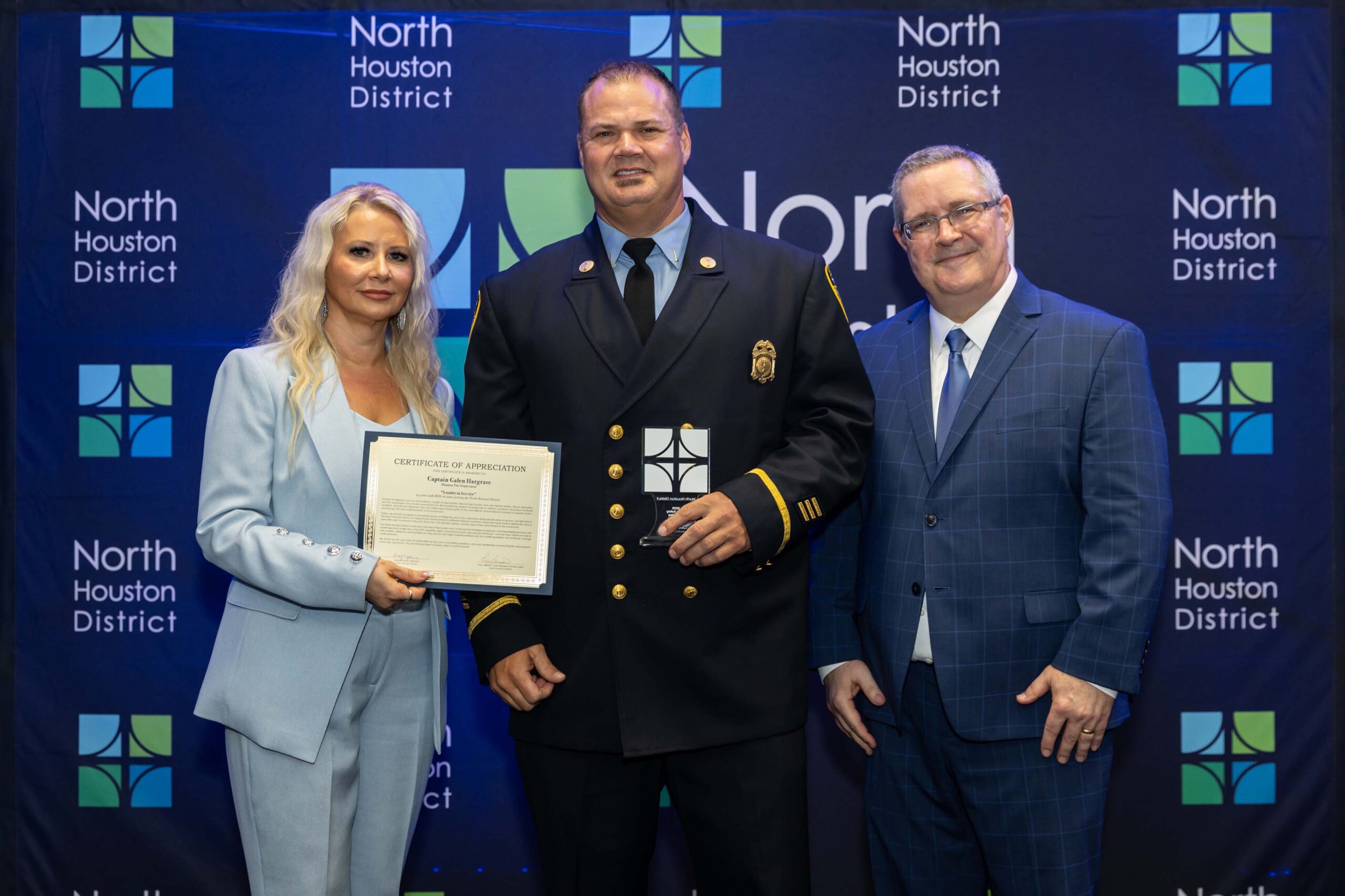 A woman in a light blue suit and a fire department captain pose on stage holding a certificate and a small award plaque. A man in a blue suit stands beside them.