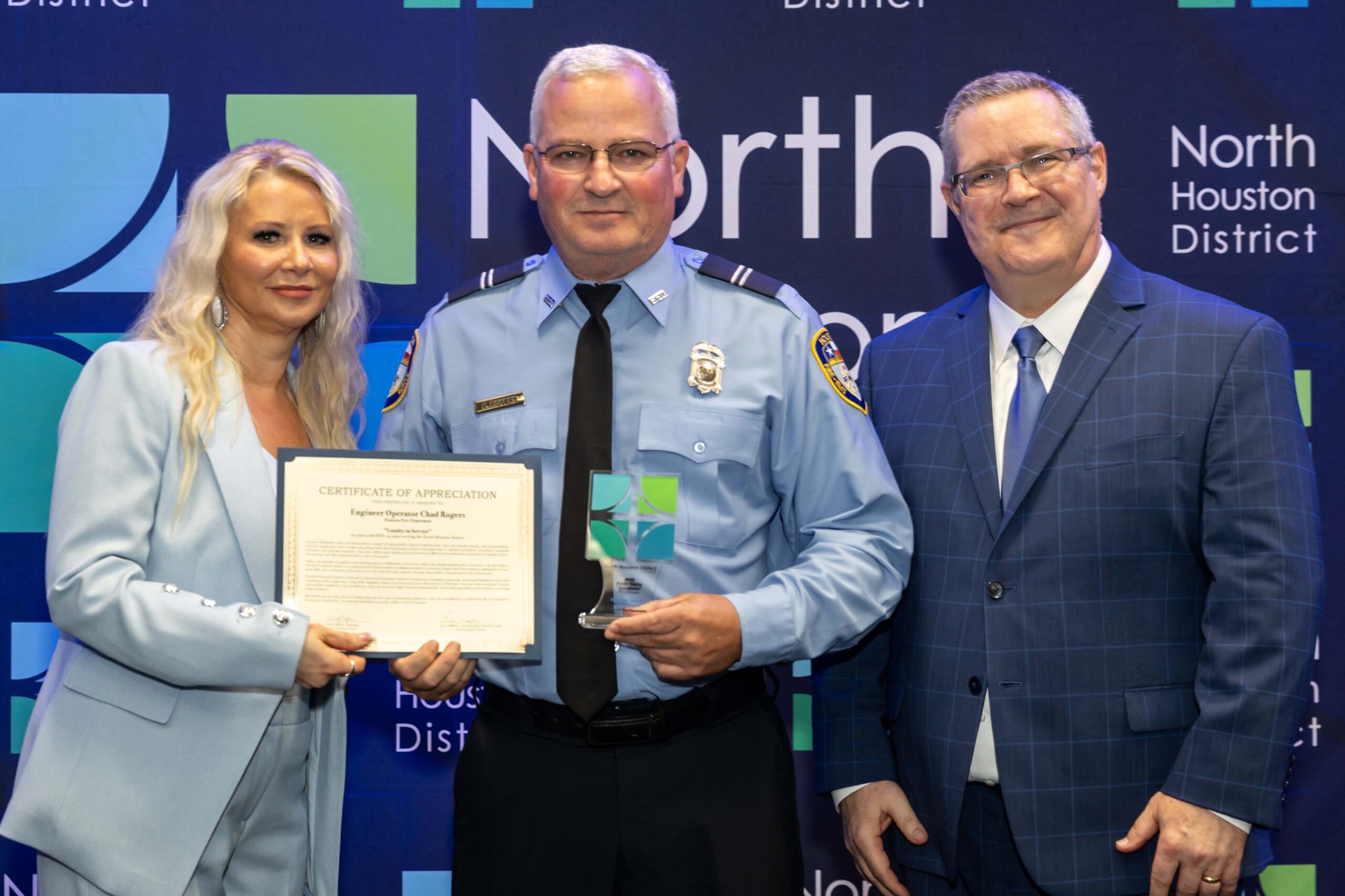 A uniformed firefighter holds a certificate and award while standing between a woman in a light blue suit and a man in a blue plaid suit.