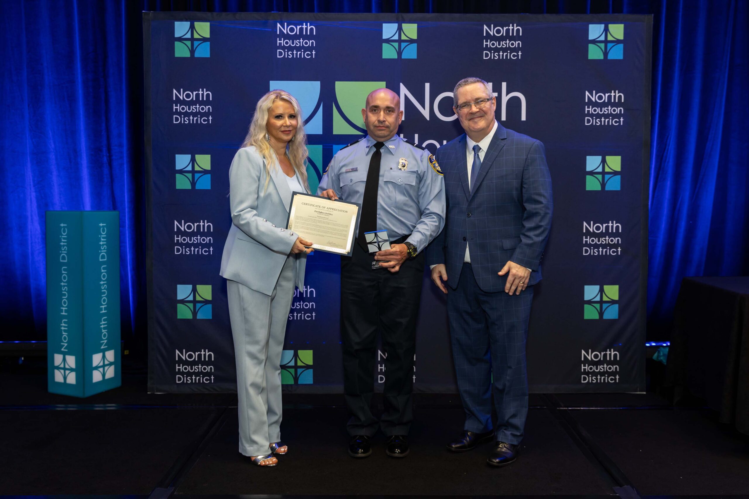 Three people stand smiling on a stage. The woman in light blue hands a certificate to a firefighter in a light blue shirt, while another man stands beside them.