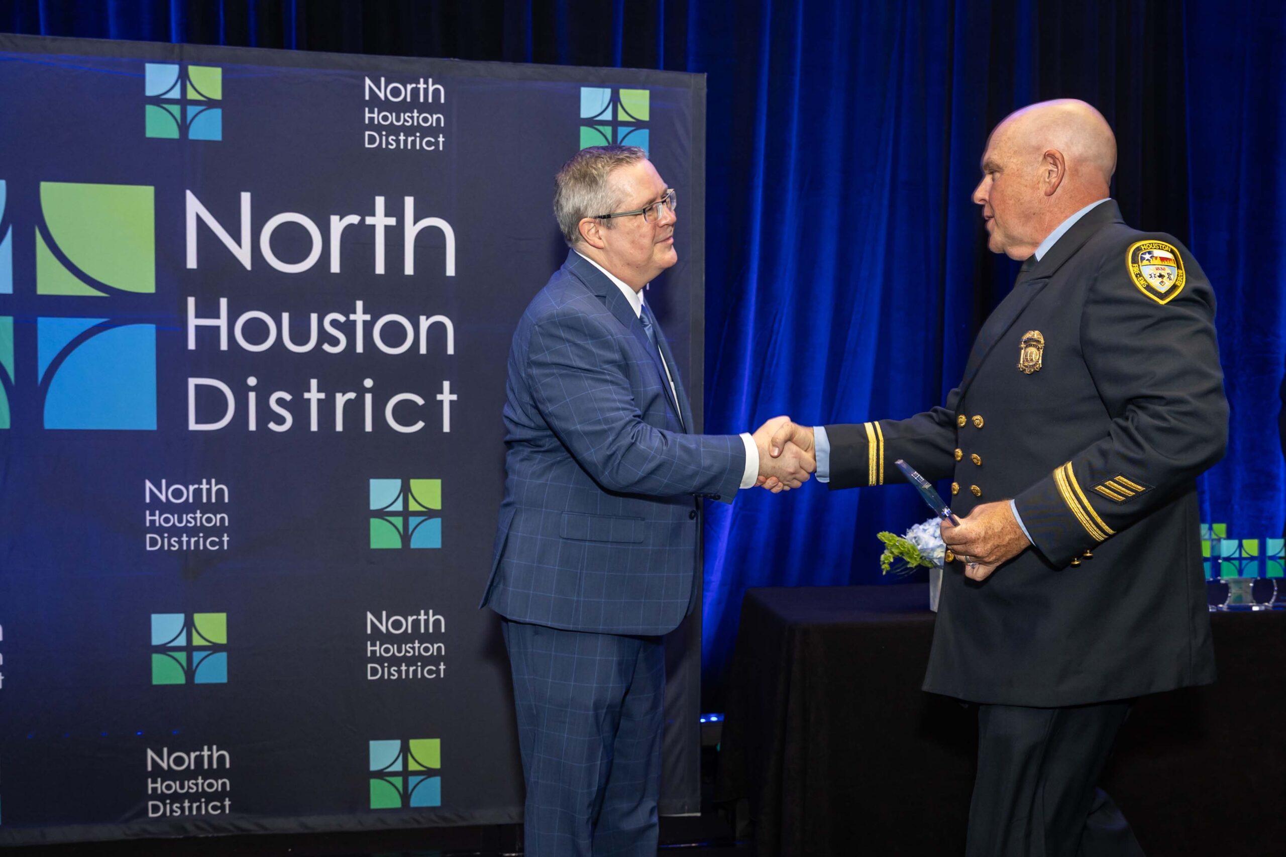 A man in a dark fire uniform shakes hands with another man in a blue plaid suit onstage in front of a branded backdrop.