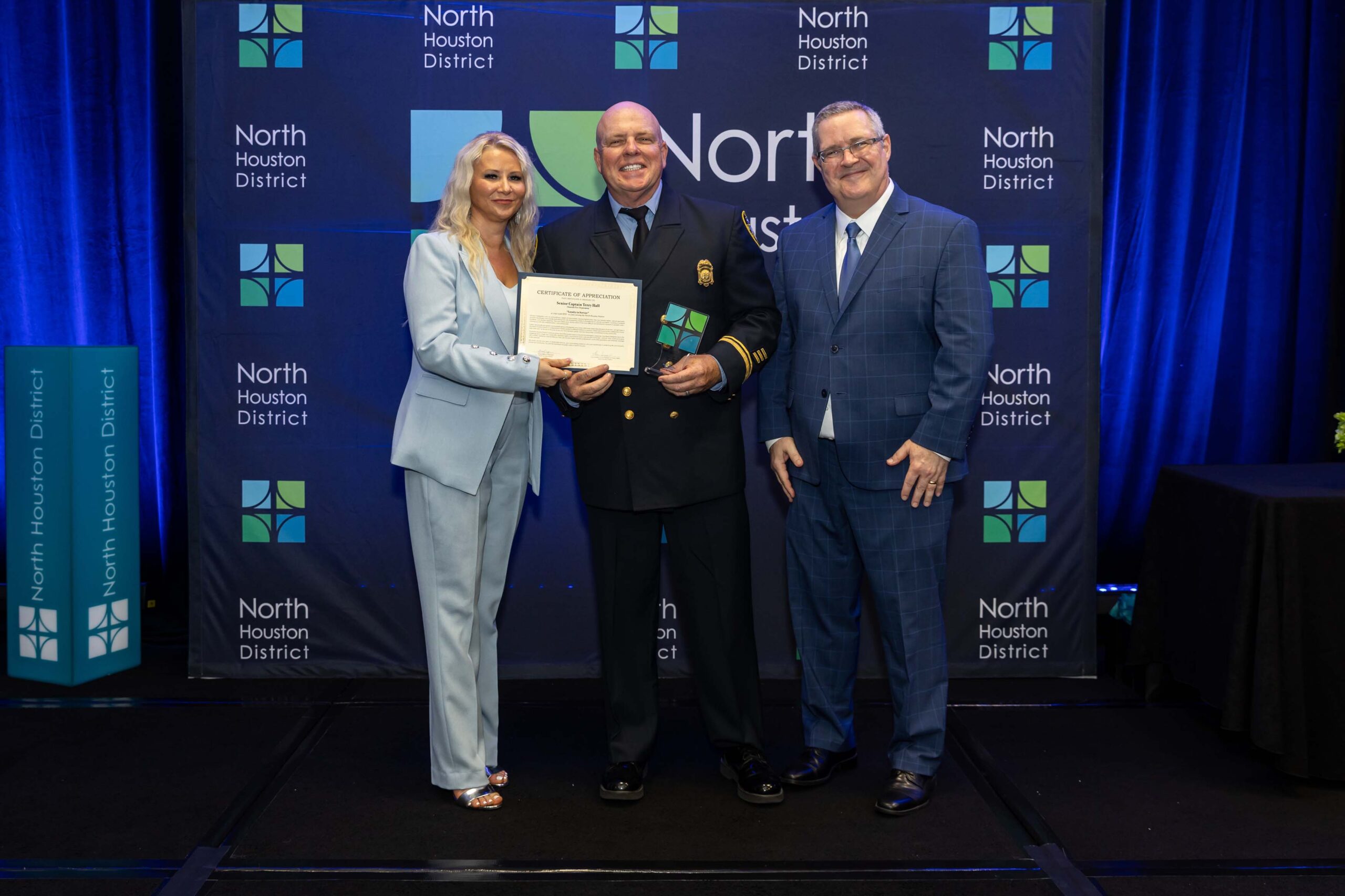 Three people stand together, smiling, in front of a North Houston District backdrop. The uniformed man holds a certificate and award.