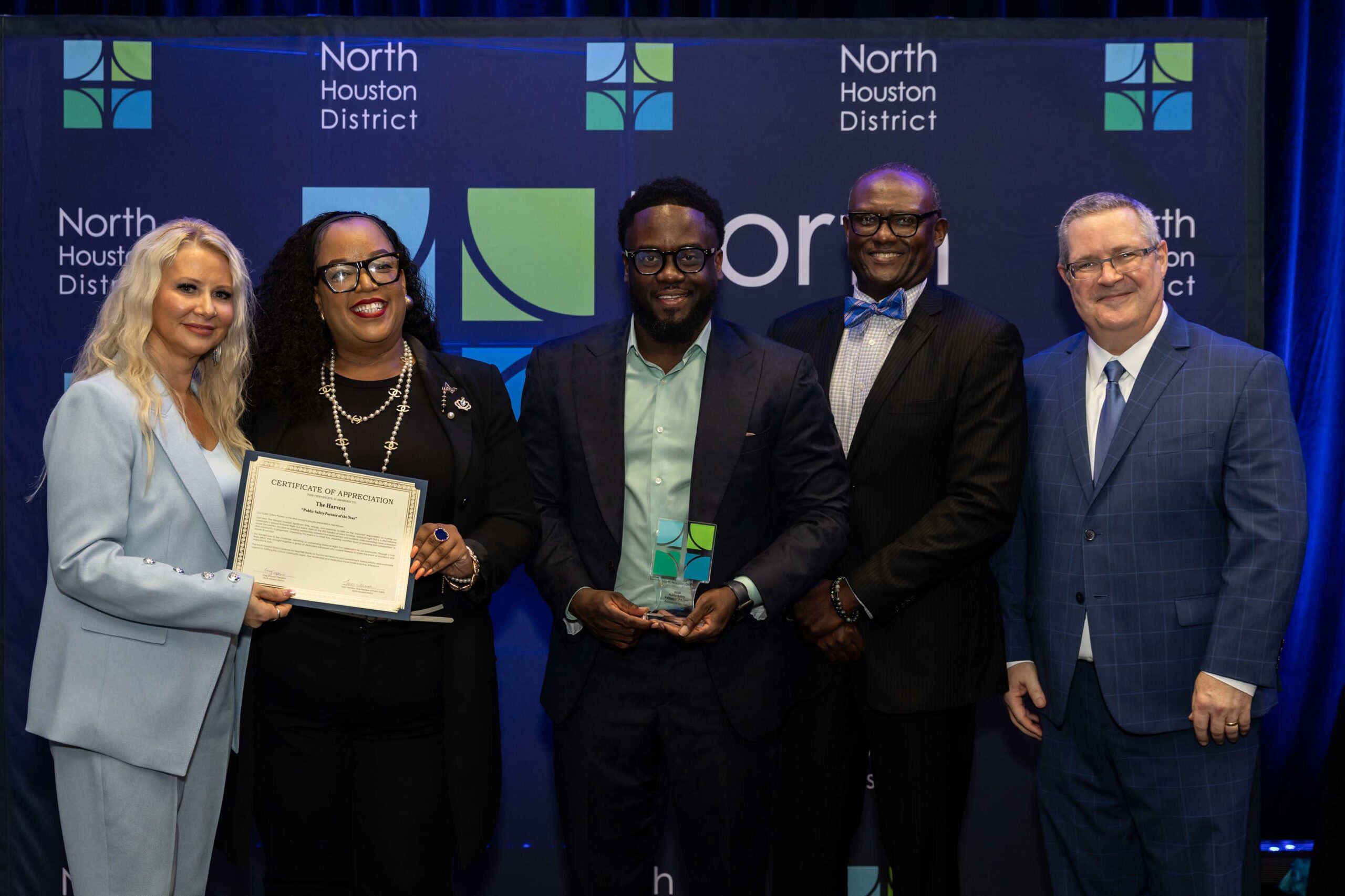 Five people pose together in front of a North Houston District backdrop. A woman in a light blue suit holds a certificate with a woman in black attire; a man in a dark suit holds a glass award.