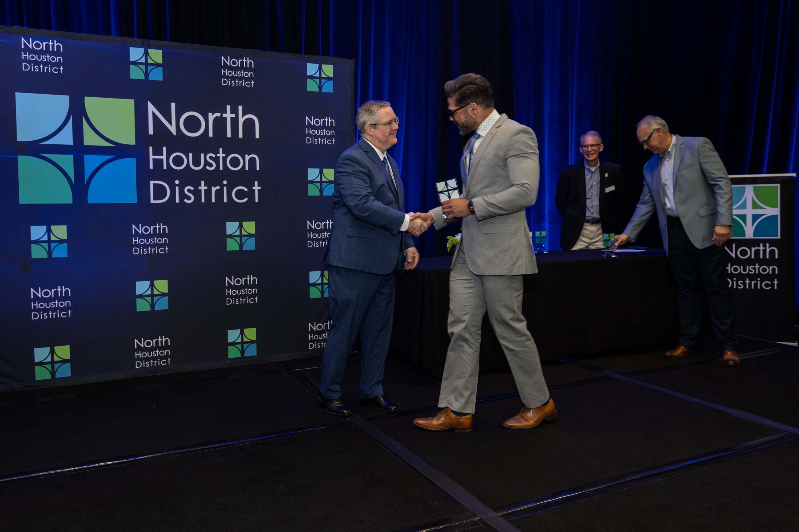 A man in a light gray suit accepts a glass award while shaking hands with a man in a blue plaid suit on stage in front of a North Houston District backdrop.