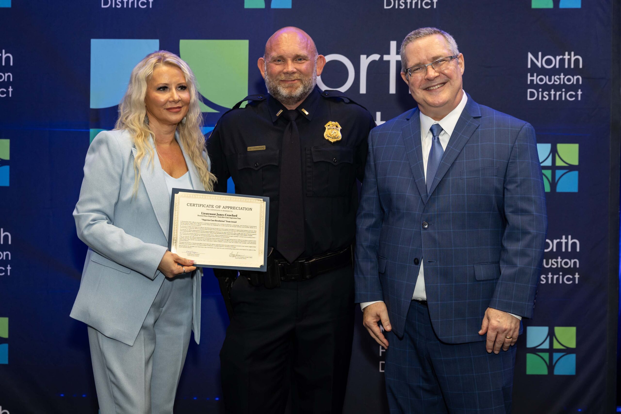 Three people stand together smiling. A woman in a blue suit holds a certificate with a police lieutenant in uniform, while a man in a blue plaid suit stands to the side.
