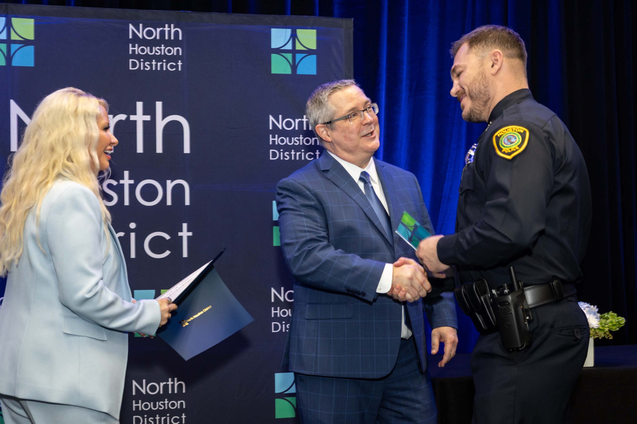 Three people stand together smiling. A woman in a blue suit holds a certificate with a police officer in uniform, while a man in a blue plaid suit stands to the side.
