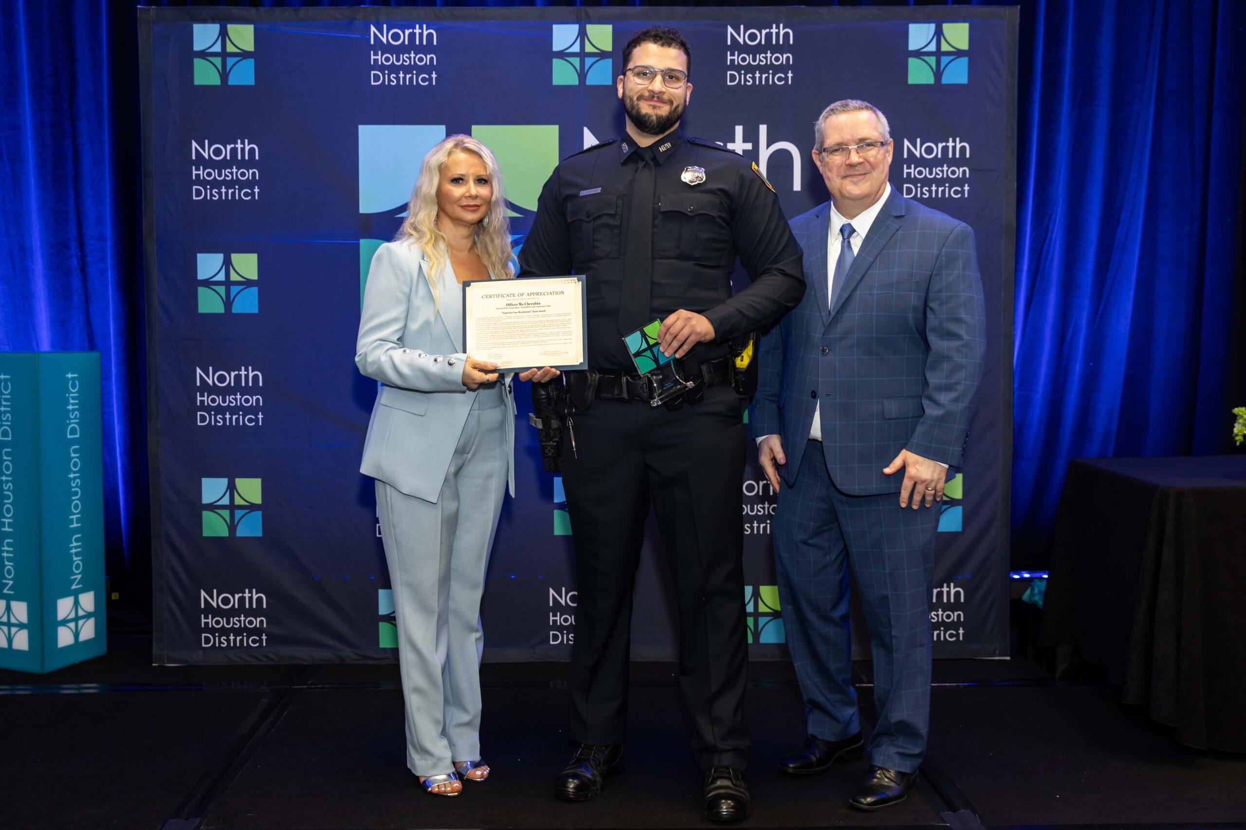 Three people stand together smiling. A woman in a blue suit holds a certificate with a police officer in uniform, while a man in a blue plaid suit stands to the side.