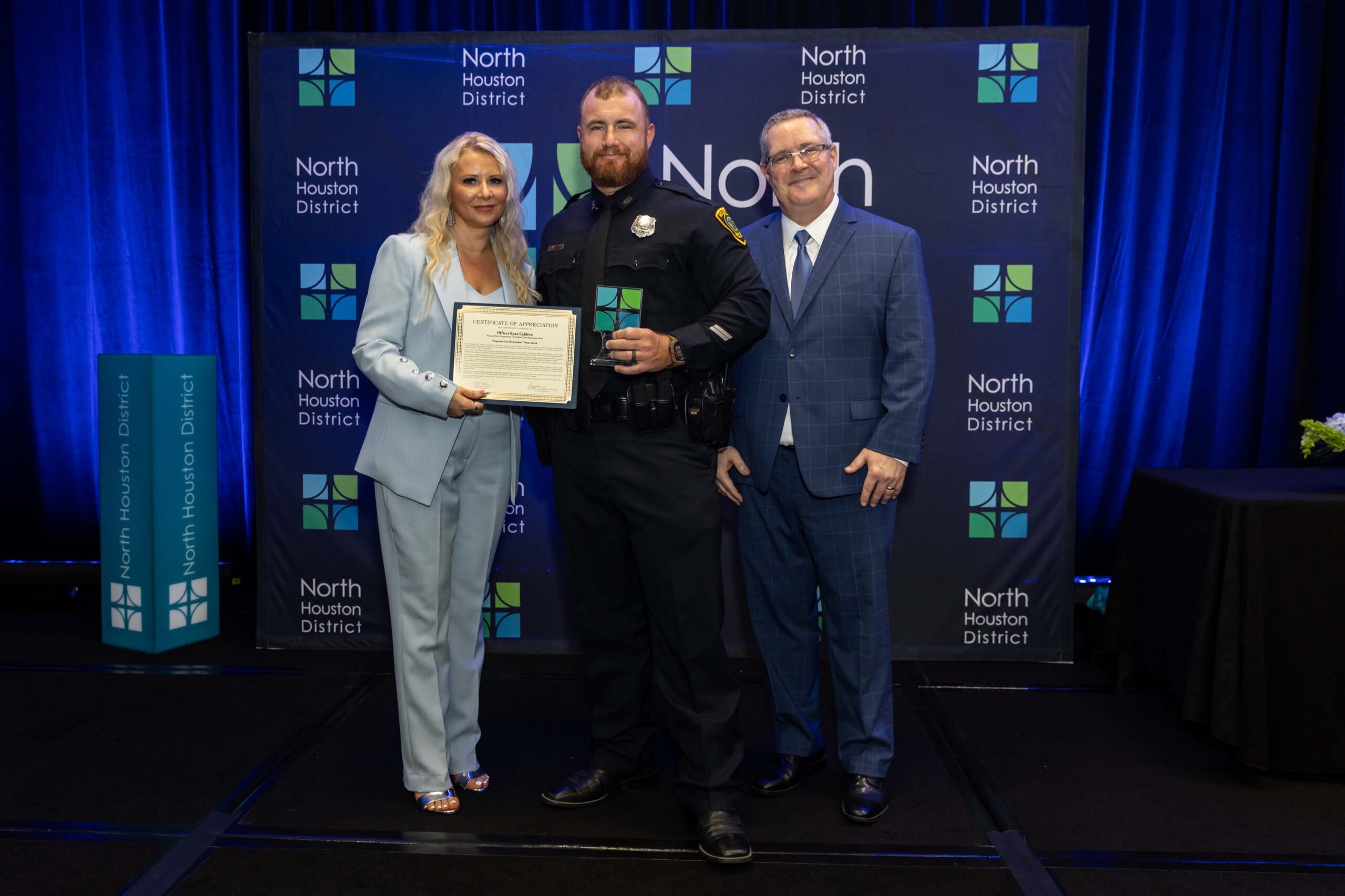 Three people stand together smiling. A woman in a blue suit holds a certificate with a police officer in uniform, while a man in a blue plaid suit stands to the side.