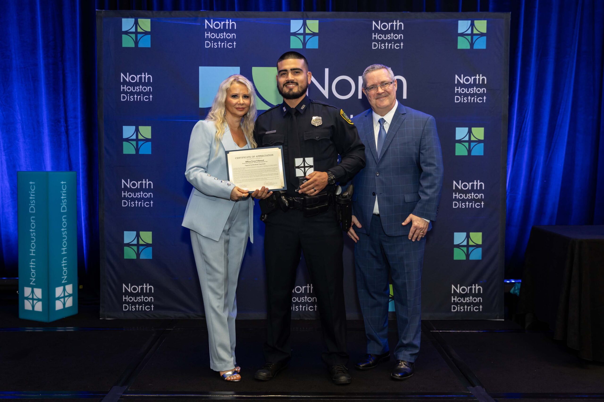 Three people stand together smiling. A woman in a blue suit holds a certificate with a police officer in uniform, while a man in a blue plaid suit stands to the side.