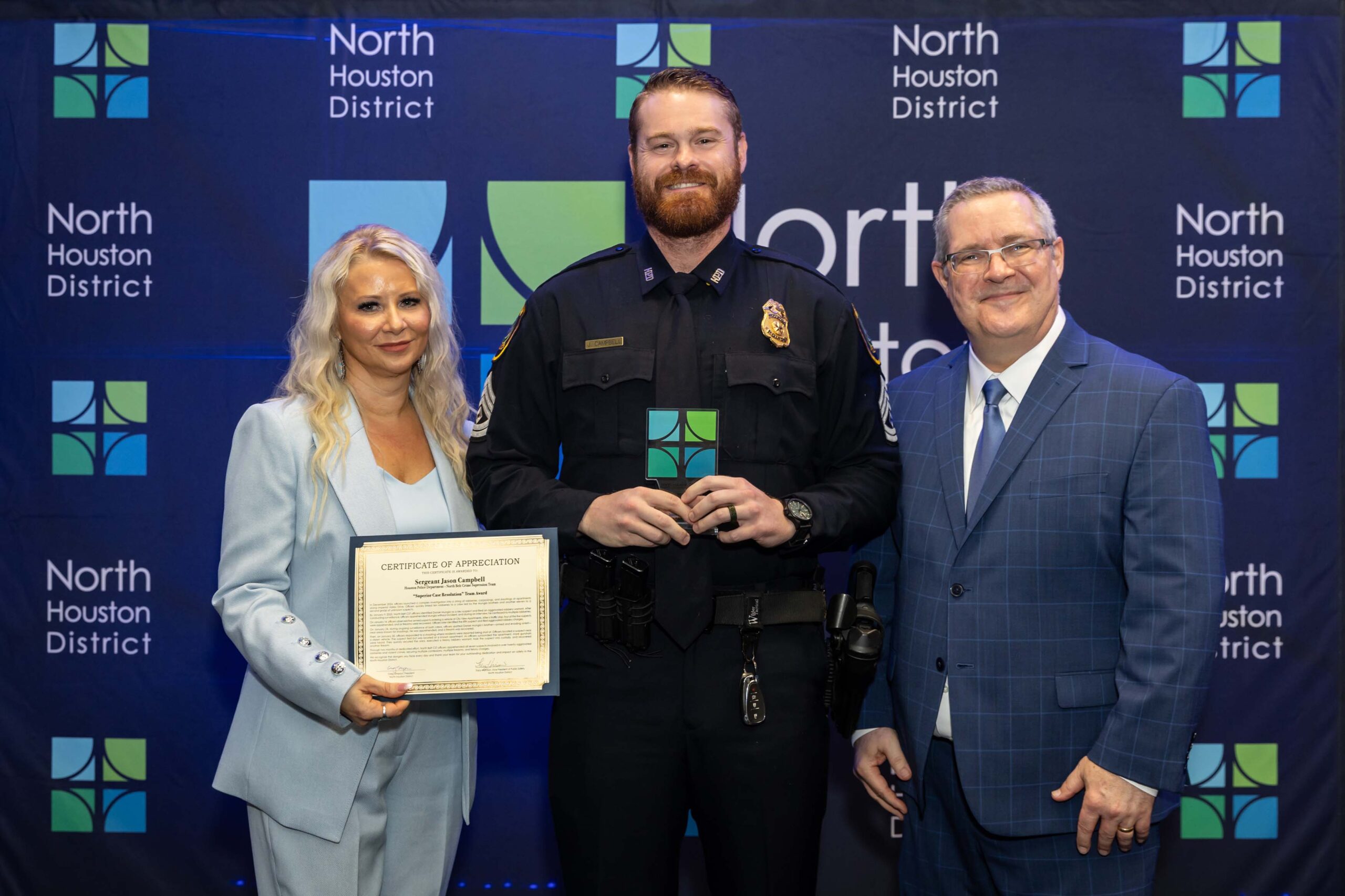 Three people stand together smiling. A woman in a blue suit holds a certificate with a police officer in uniform, while a man in a blue plaid suit stands to the side.
