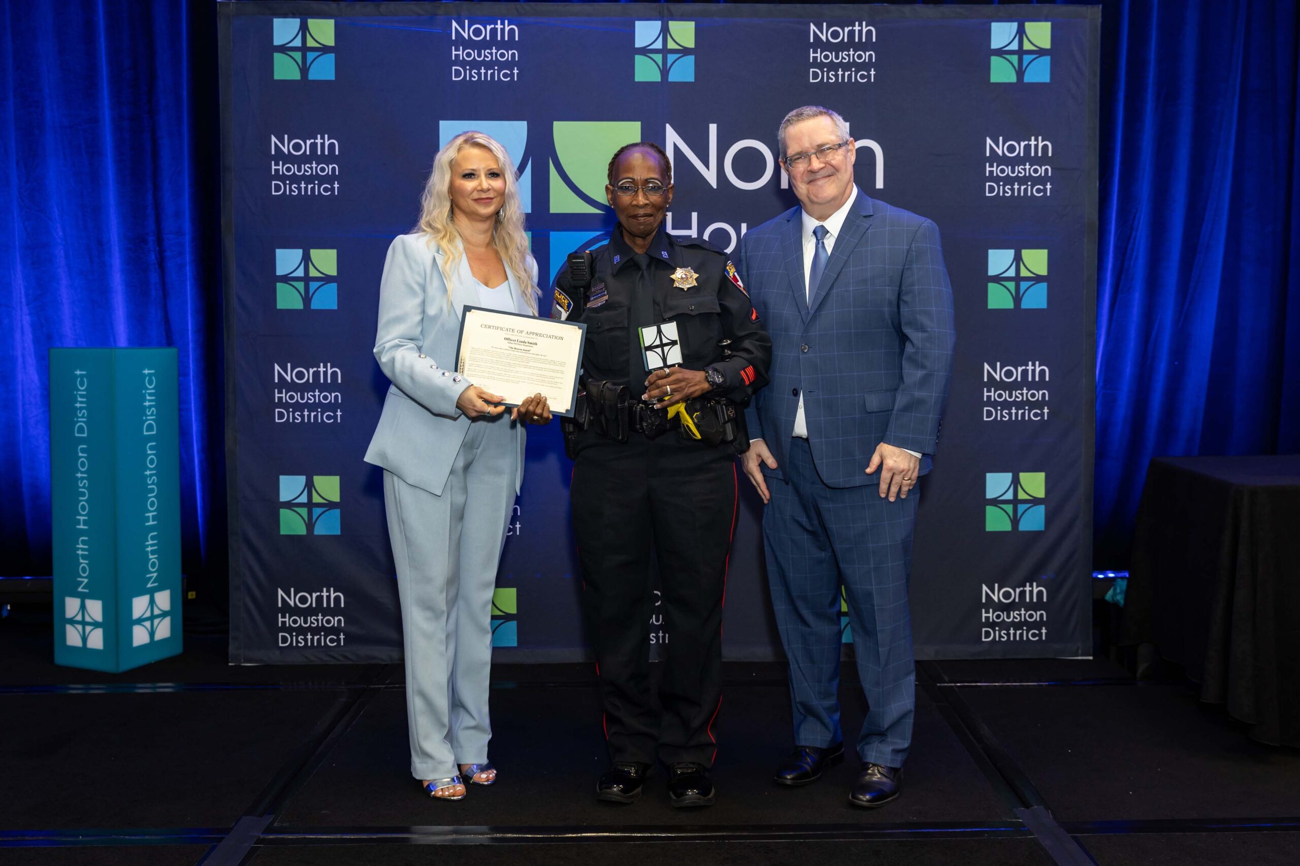 Three people stand together smiling. A woman in a blue suit holds a certificate with a police officer in uniform, while a man in a blue plaid suit stands to the side.