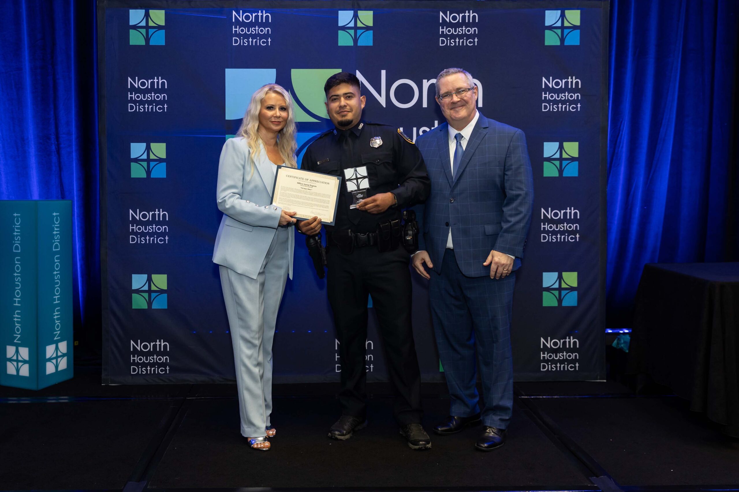 Three people stand together smiling. A woman in a blue suit holds a certificate with a police officer in uniform, while a man in a blue plaid suit stands to the side.