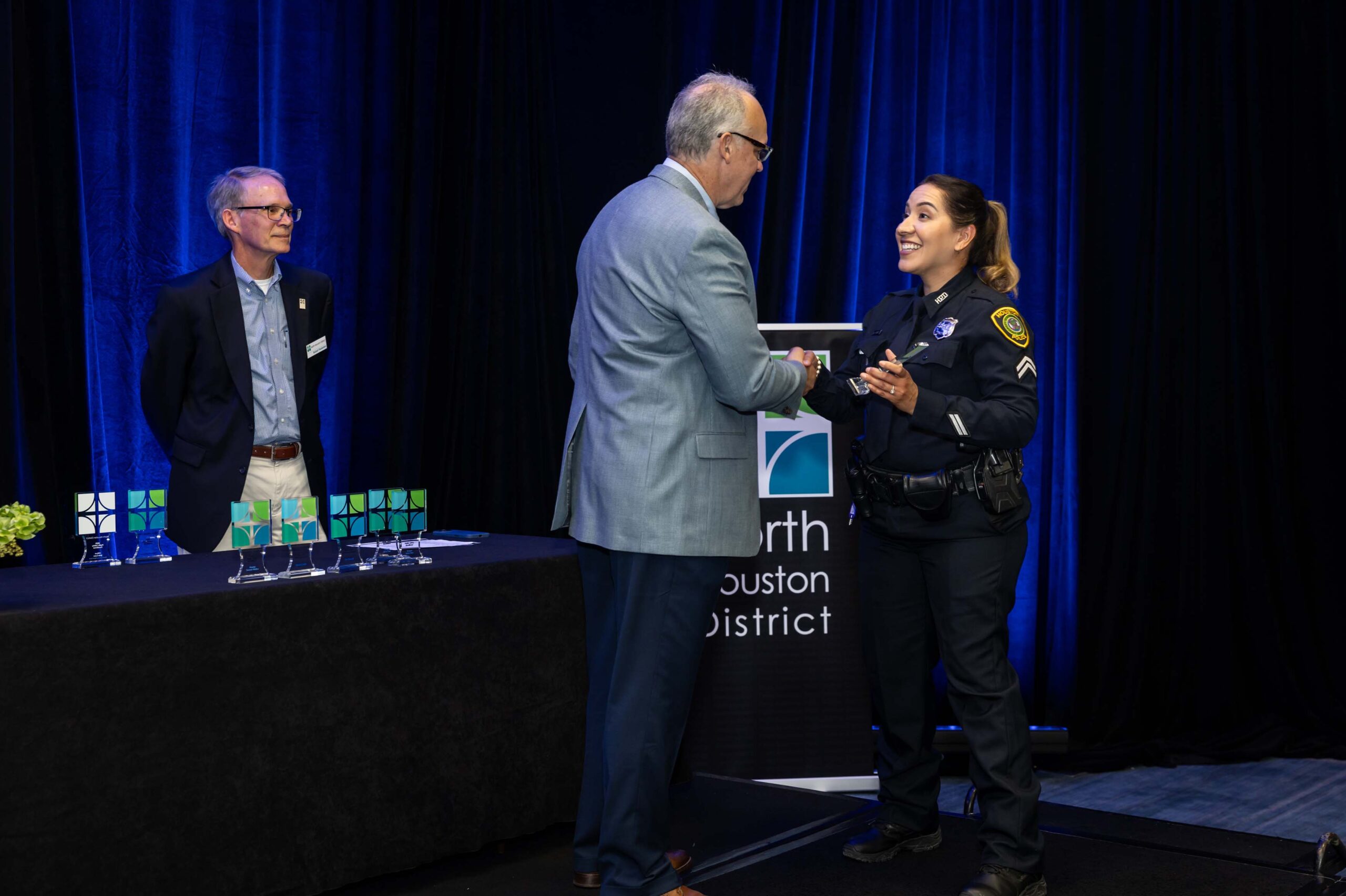 A uniformed officer receives her award while smiling and shaking hands with a presenter on stage during the Public Safety Awards, with trophies visible on the table behind them.