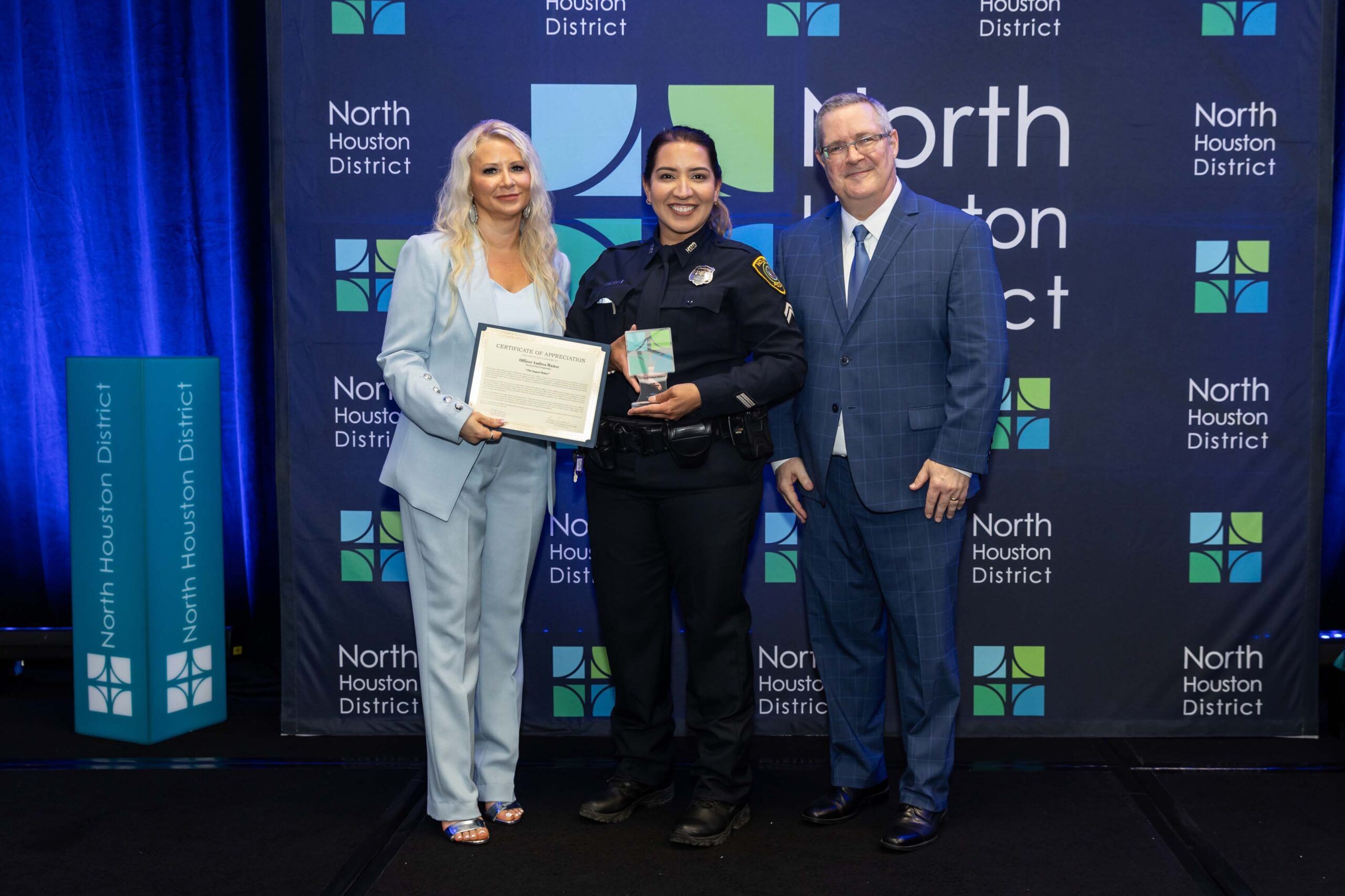 Three people stand together smiling. A woman in a blue suit holds a certificate with a police officer in uniform, while a man in a blue plaid suit stands to the side.